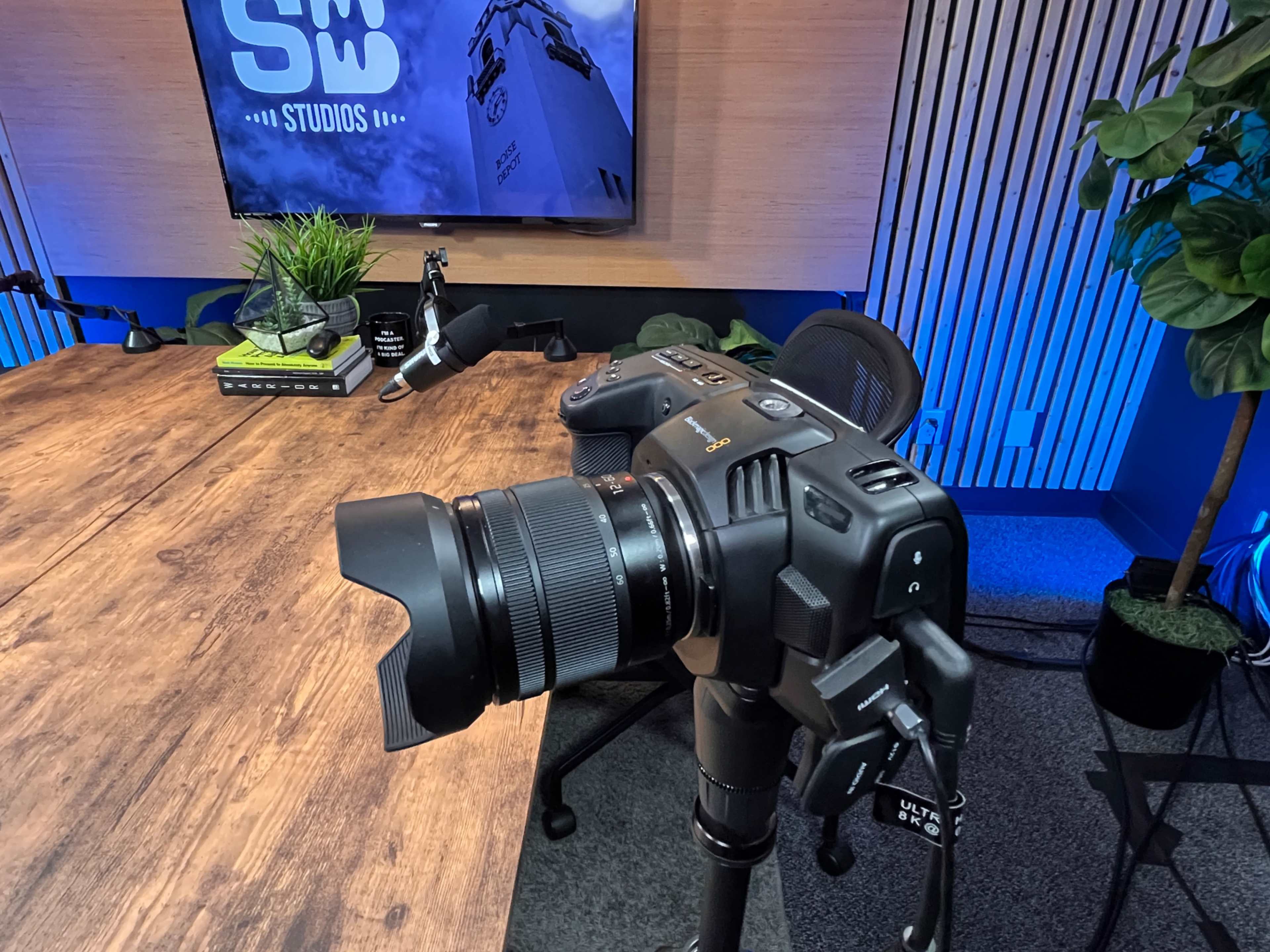 A professional camera is positioned on a tripod in a studio setting with a wooden table and a large screen displaying the text "SW STUDIOS" in the background.