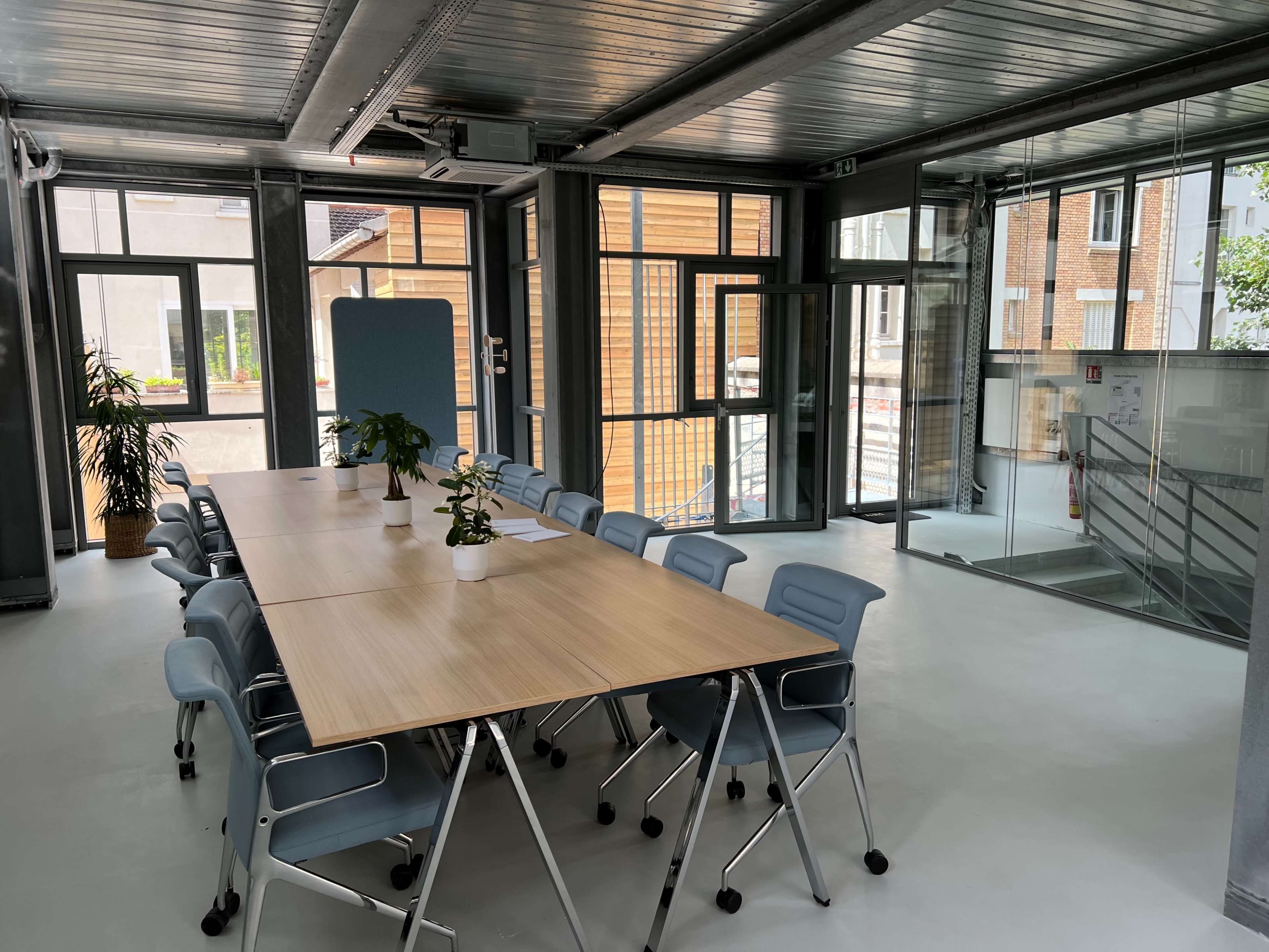 A modern conference room features a long wooden table surrounded by light blue chairs, with large windows providing natural light.