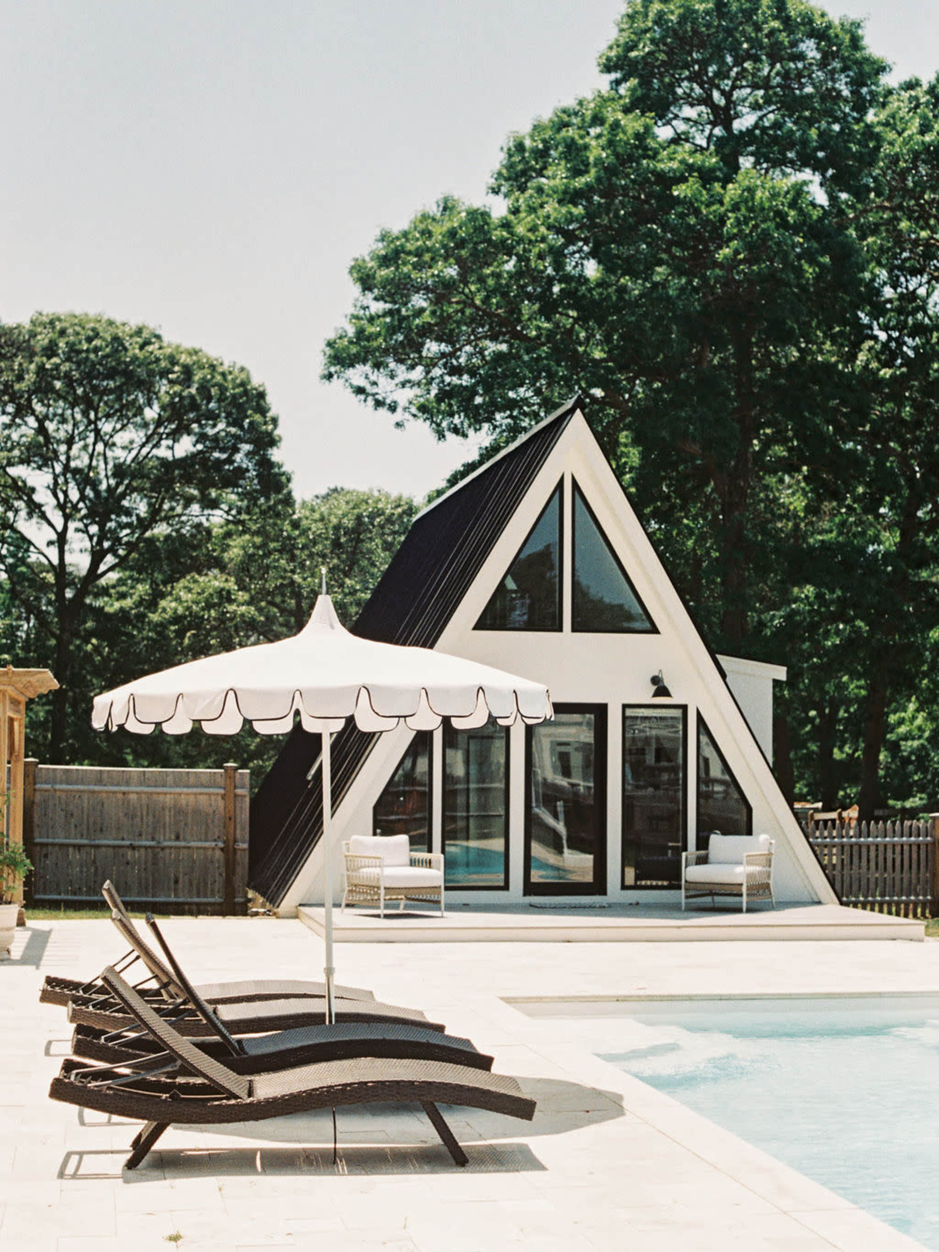 A modern A-frame house with large glass doors stands beside a swimming pool adorned with lounge chairs and an umbrella, surrounded by trees.