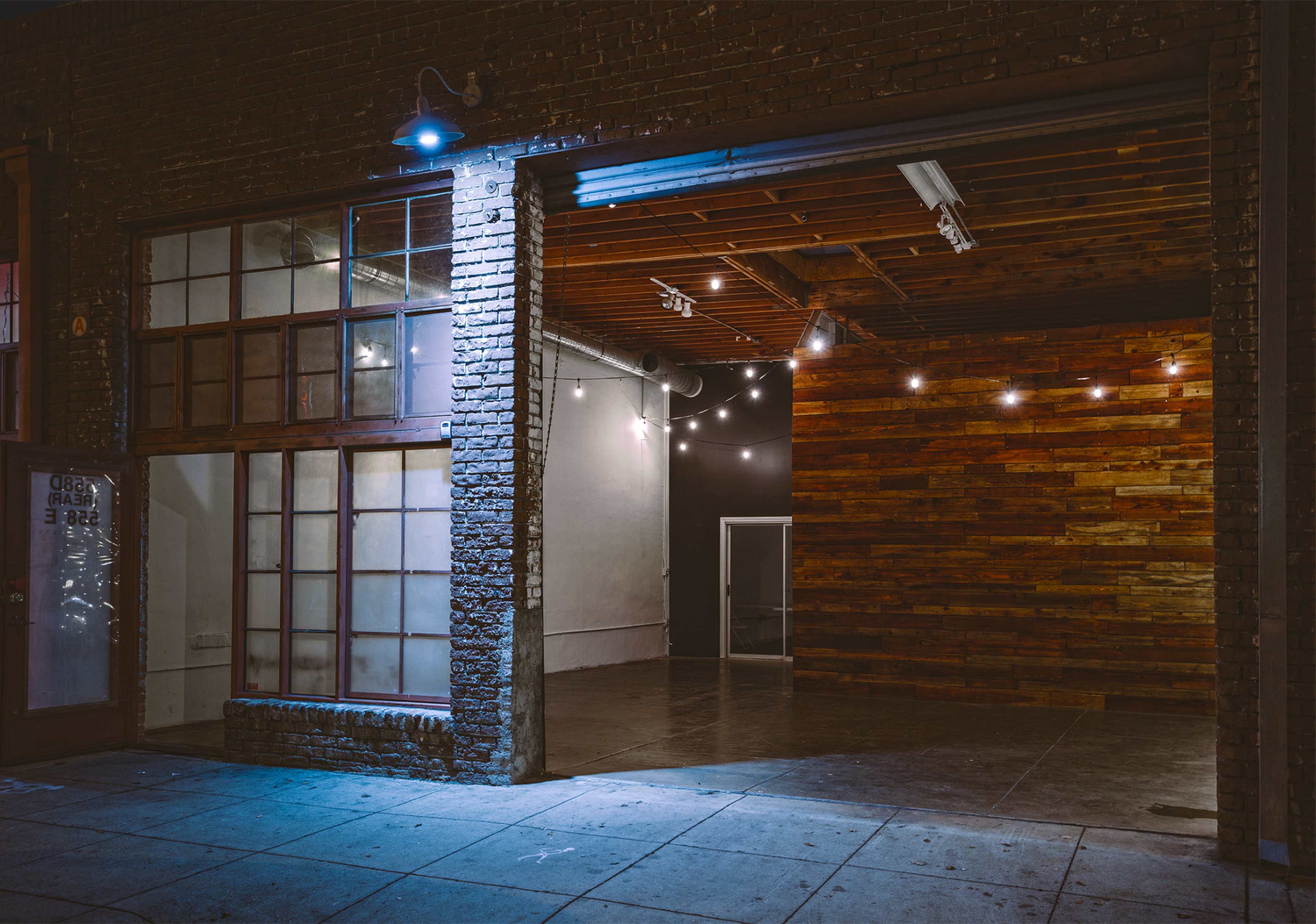 A brick building with large windows and an open entryway, revealing a sparsely furnished interior featuring wooden paneling and string lights.