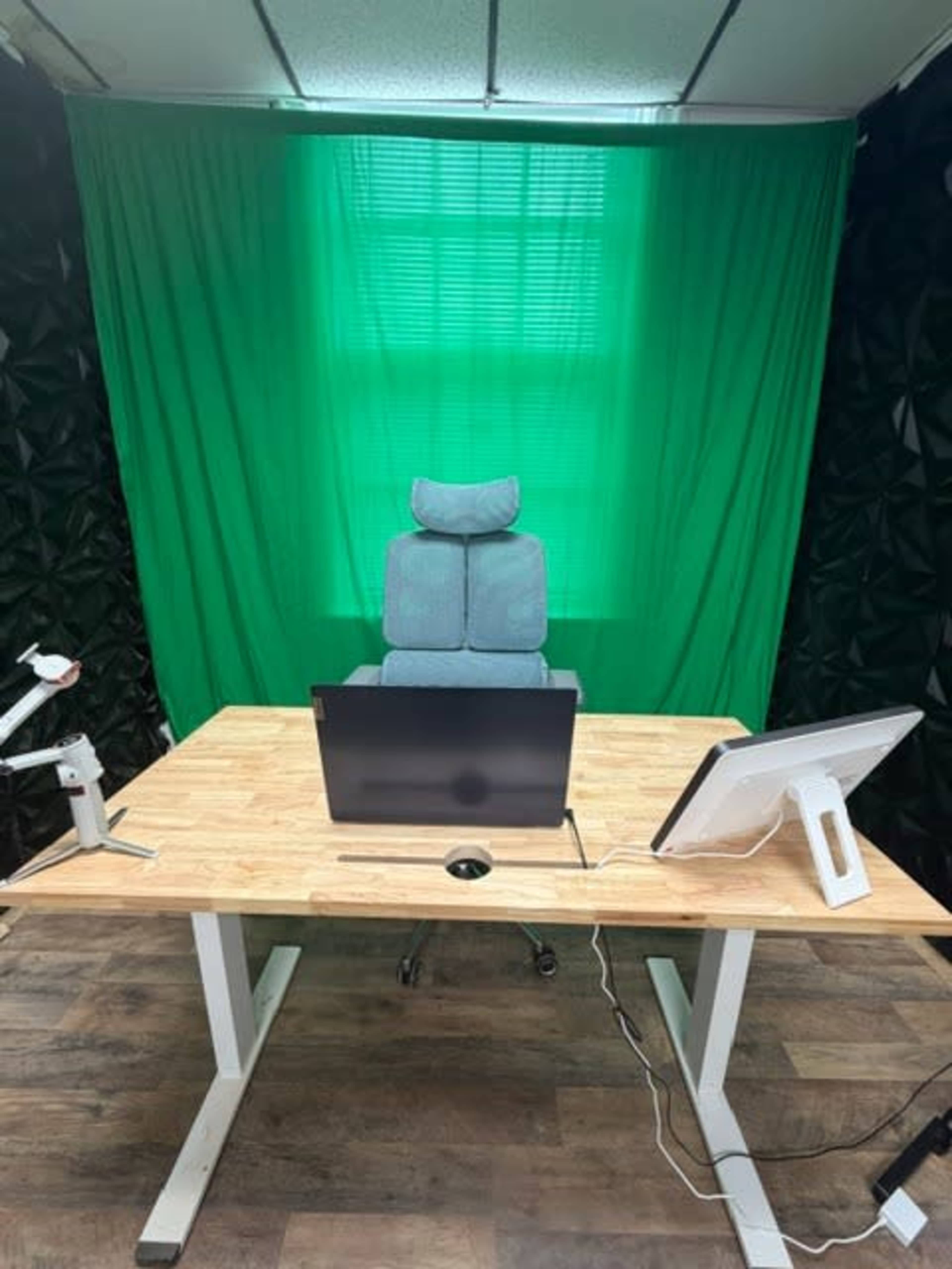 The image shows a minimalist office setup featuring a wooden desk with two monitors, an ergonomic chair in front of a green backdrop, and a wall with a textured design.
