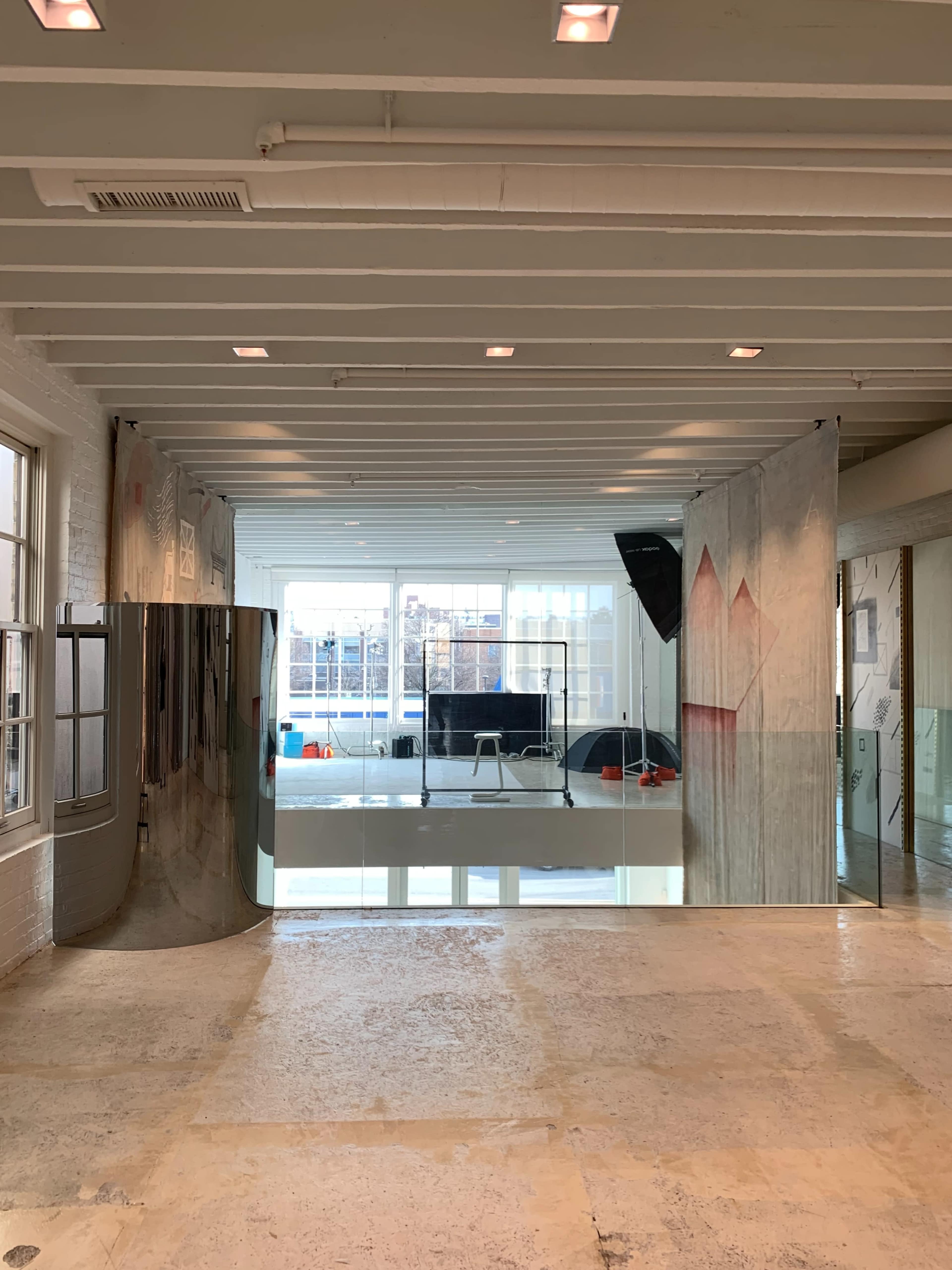 The image shows an interior space with large glass panels reflecting light, revealing a monitor in the background and artwork on the walls.