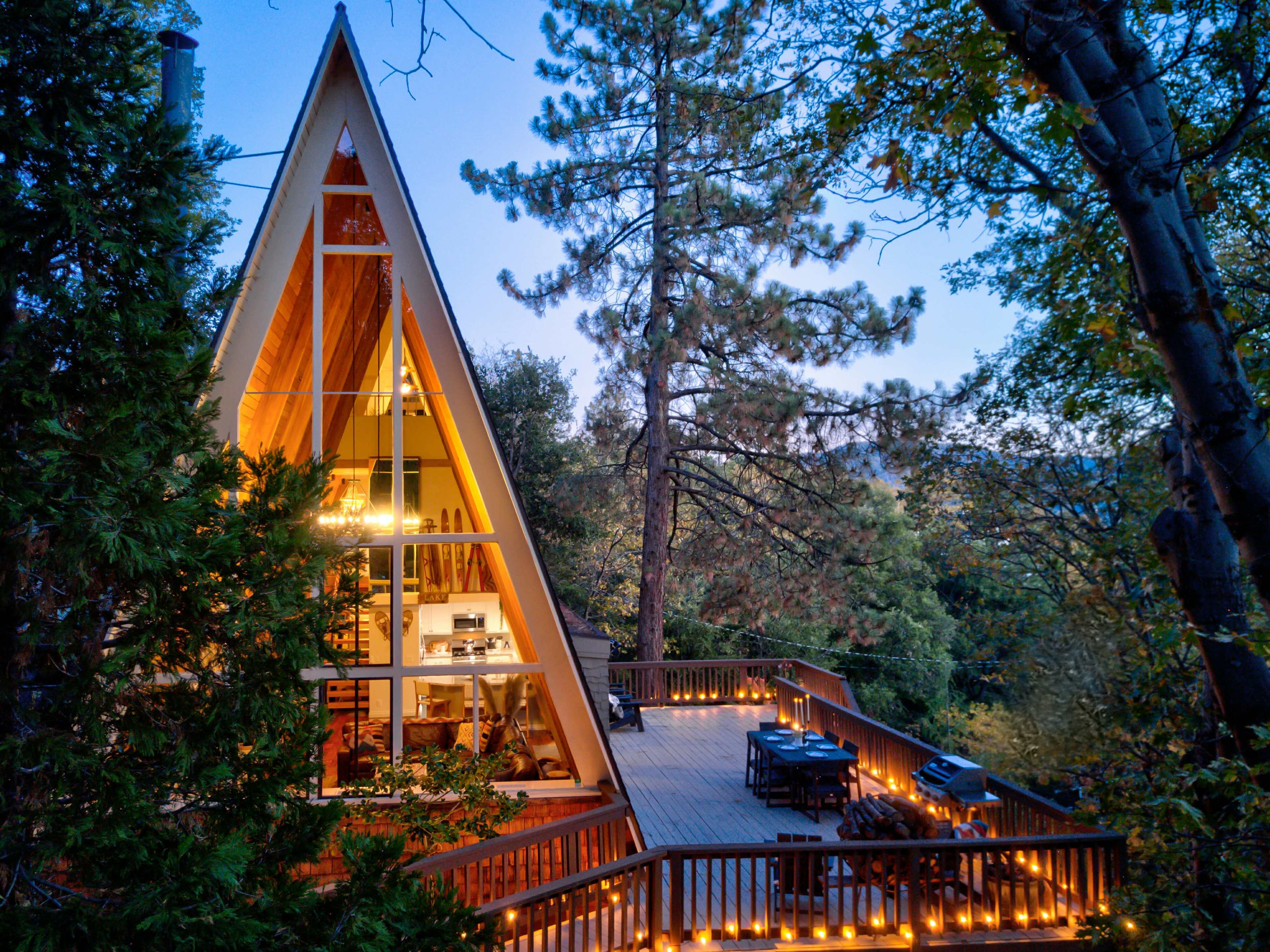 A modern A-frame cabin surrounded by trees, featuring large windows and a spacious wooden deck illuminated with string lights.