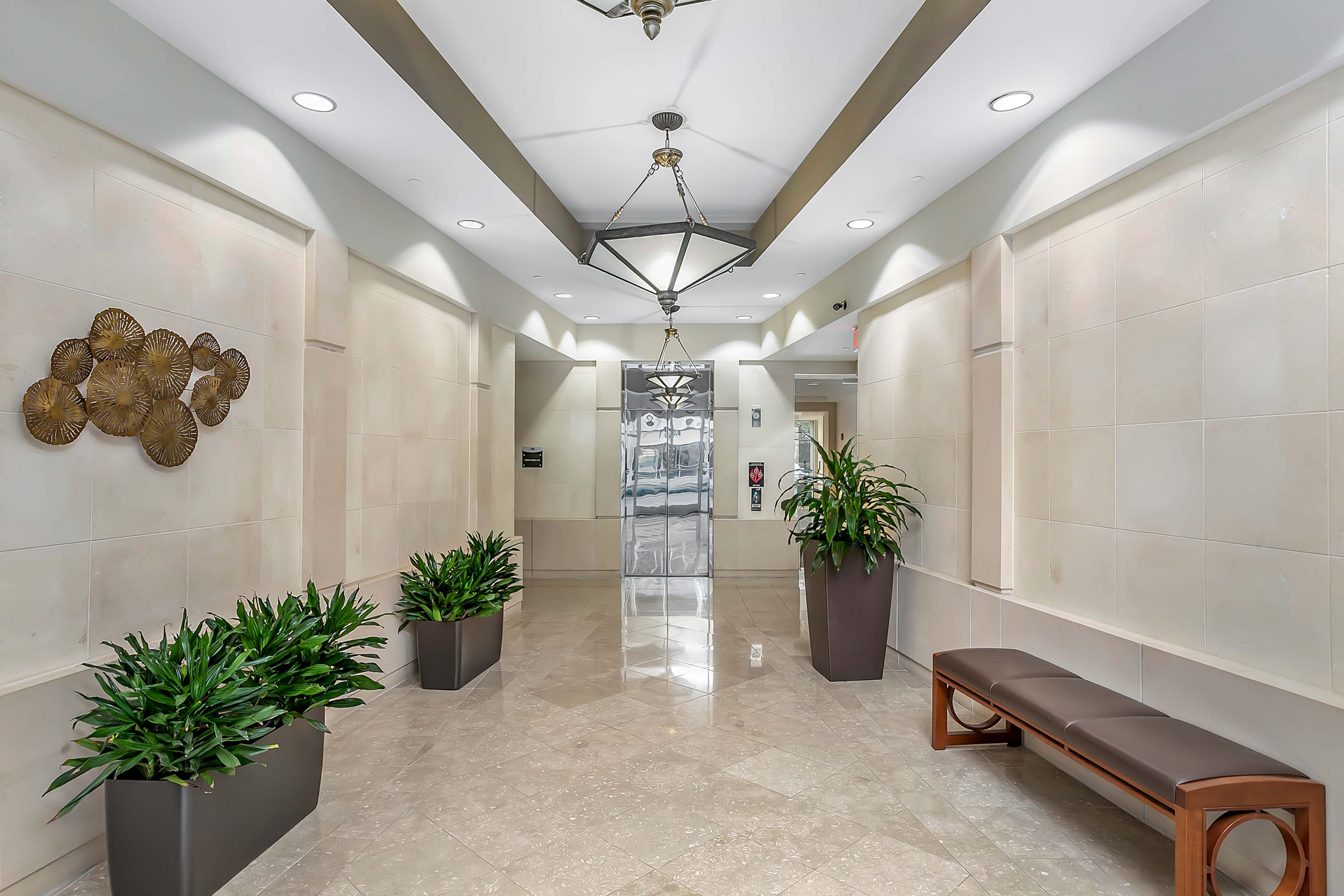 The image shows a modern lobby with a polished tile floor, plants in planters, a wooden bench, and decorative wall art.