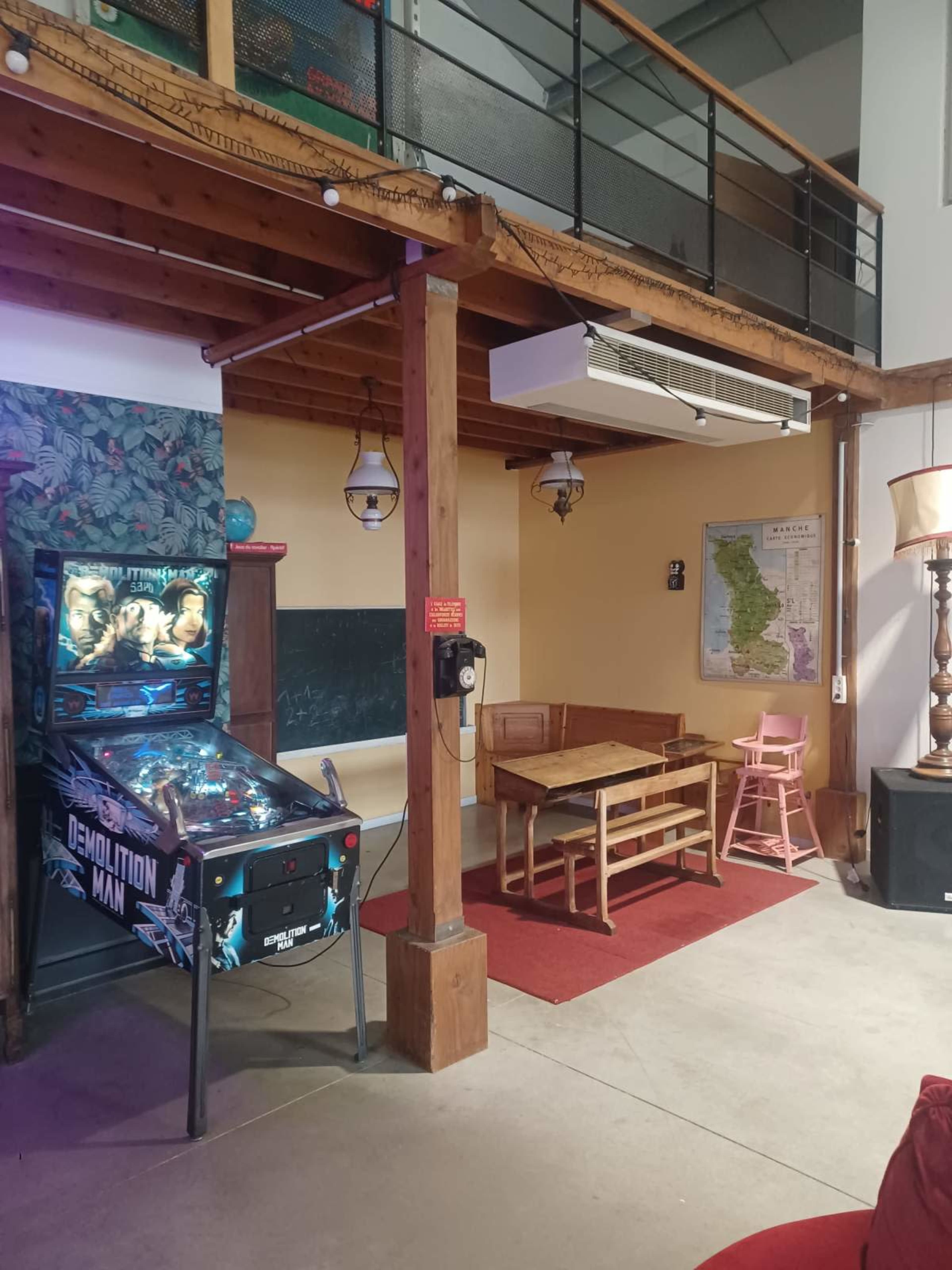 The image shows an interior space featuring a pinball machine, a dining table with chairs, and decorative elements like a map and plants against wooden beams.