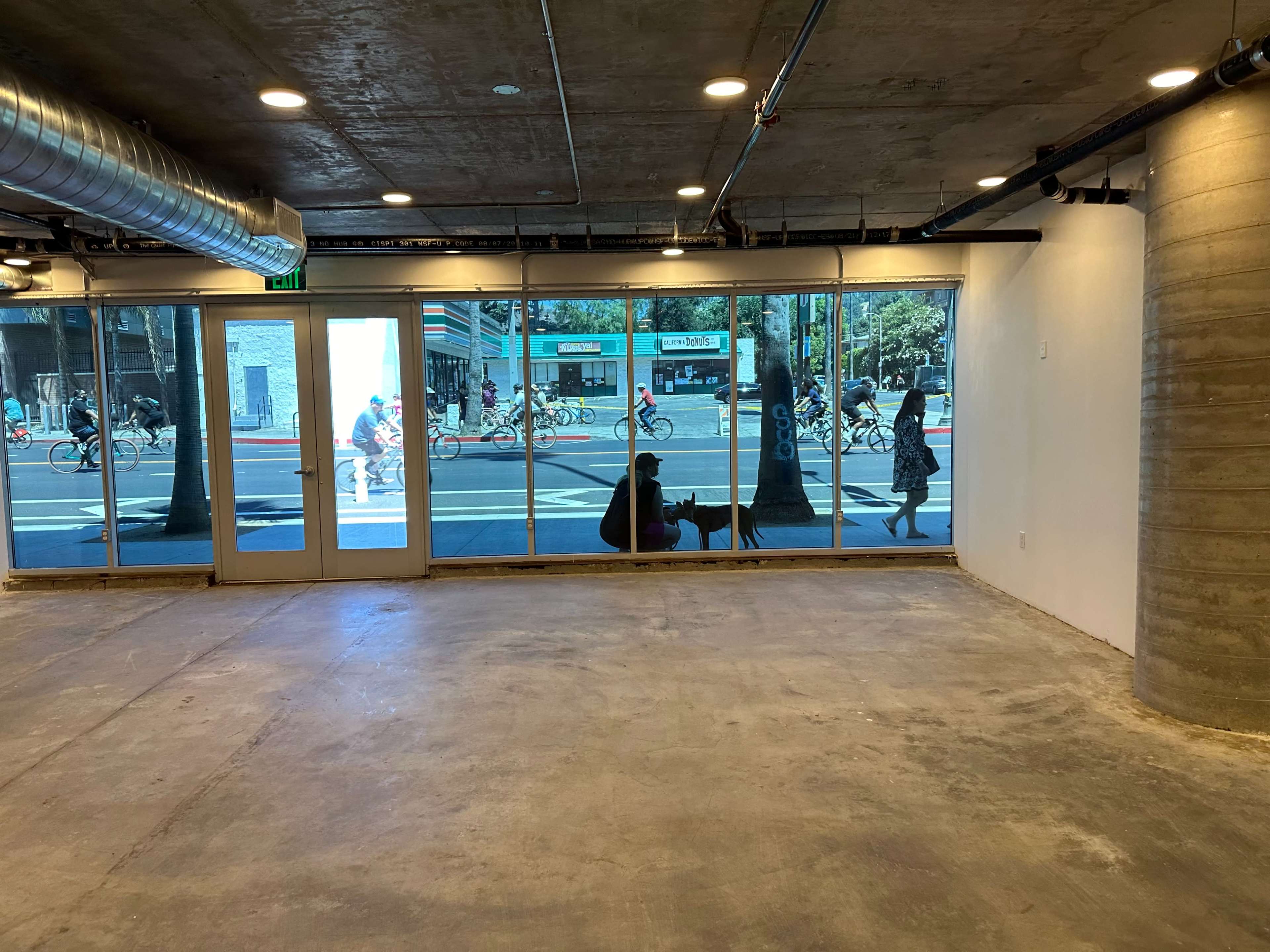 The image shows an empty interior space with large windows displaying a street scene where people and cyclists are passing by.