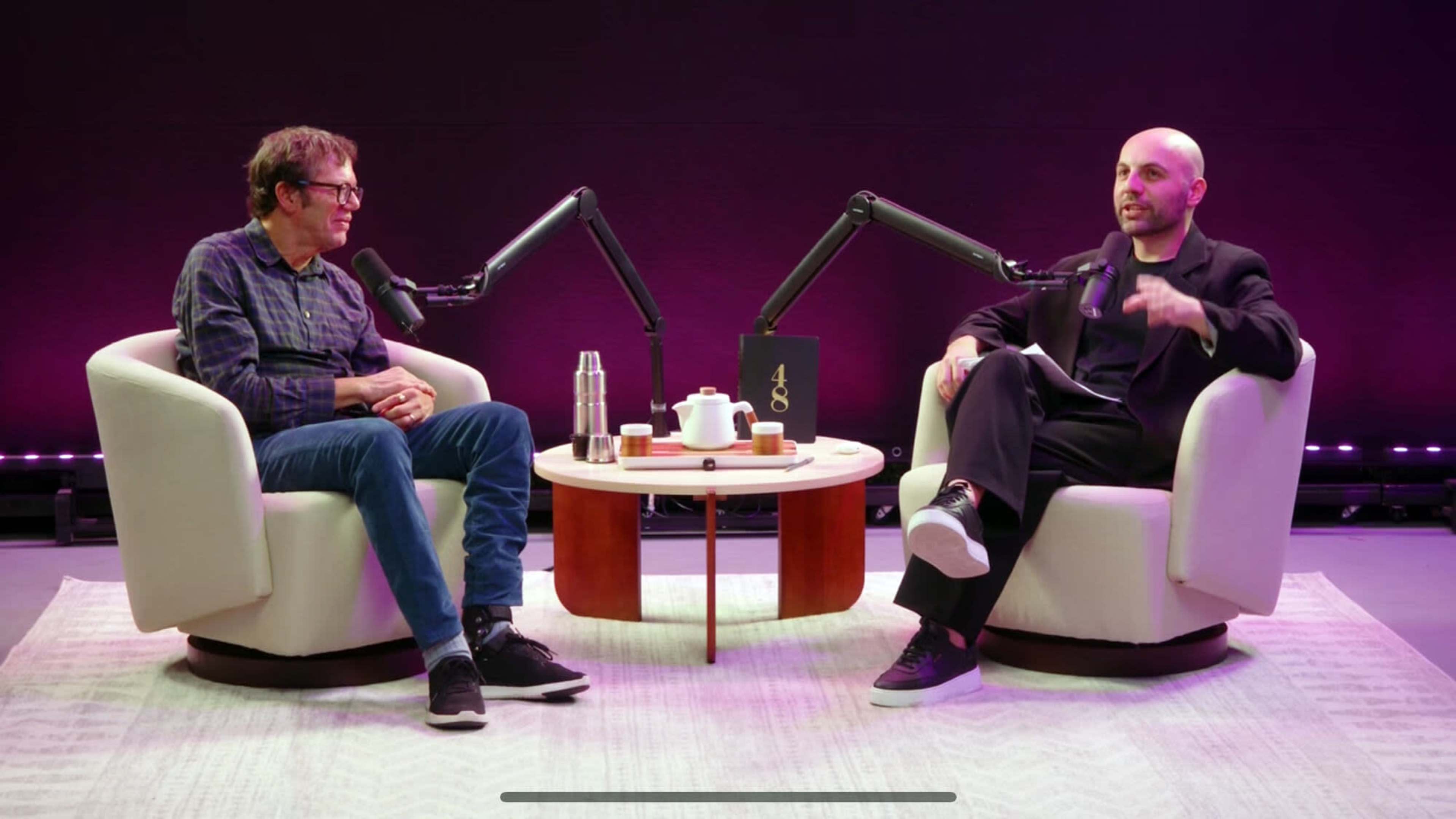 Two men sit in modern chairs with a small round table between them, equipped with podcast microphones, in front of a purple backdrop.
