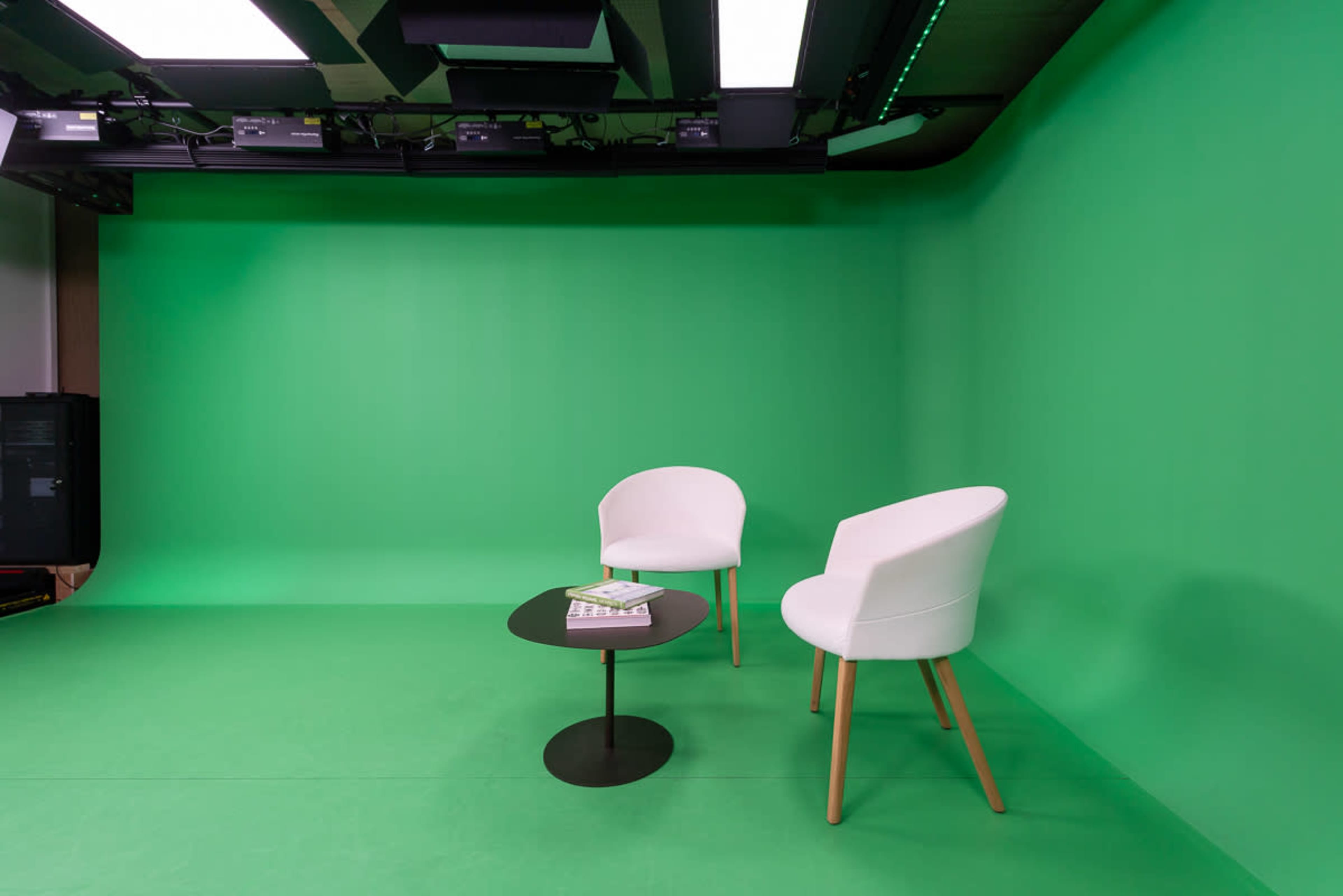 The image shows a studio space with a green screen backdrop, featuring two white chairs and a small black table.