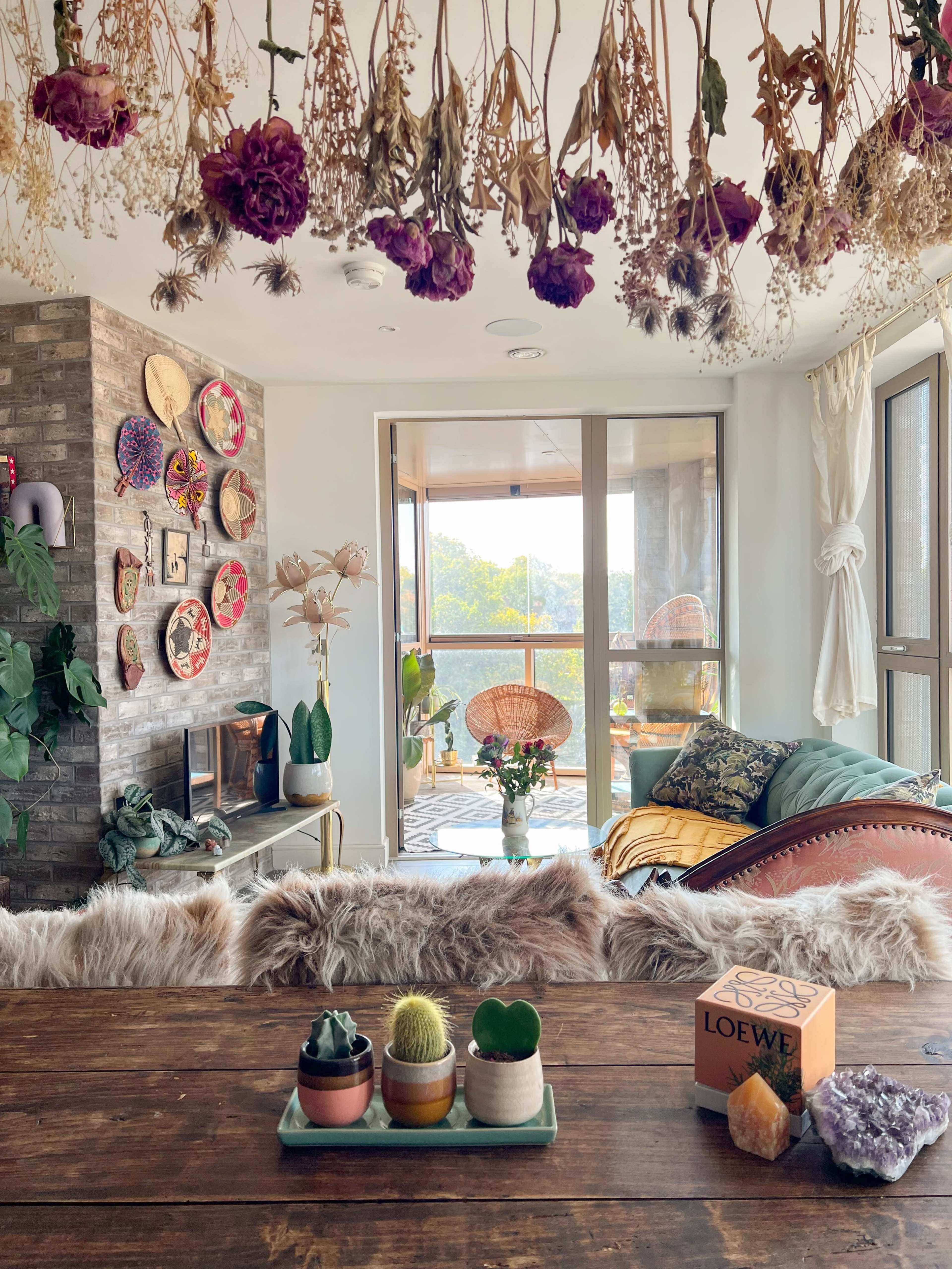 A cozy living room features a wall of dried flowers, decorative plates, and a wooden table with succulent plants, overlooking a scenic view through large glass windows.
