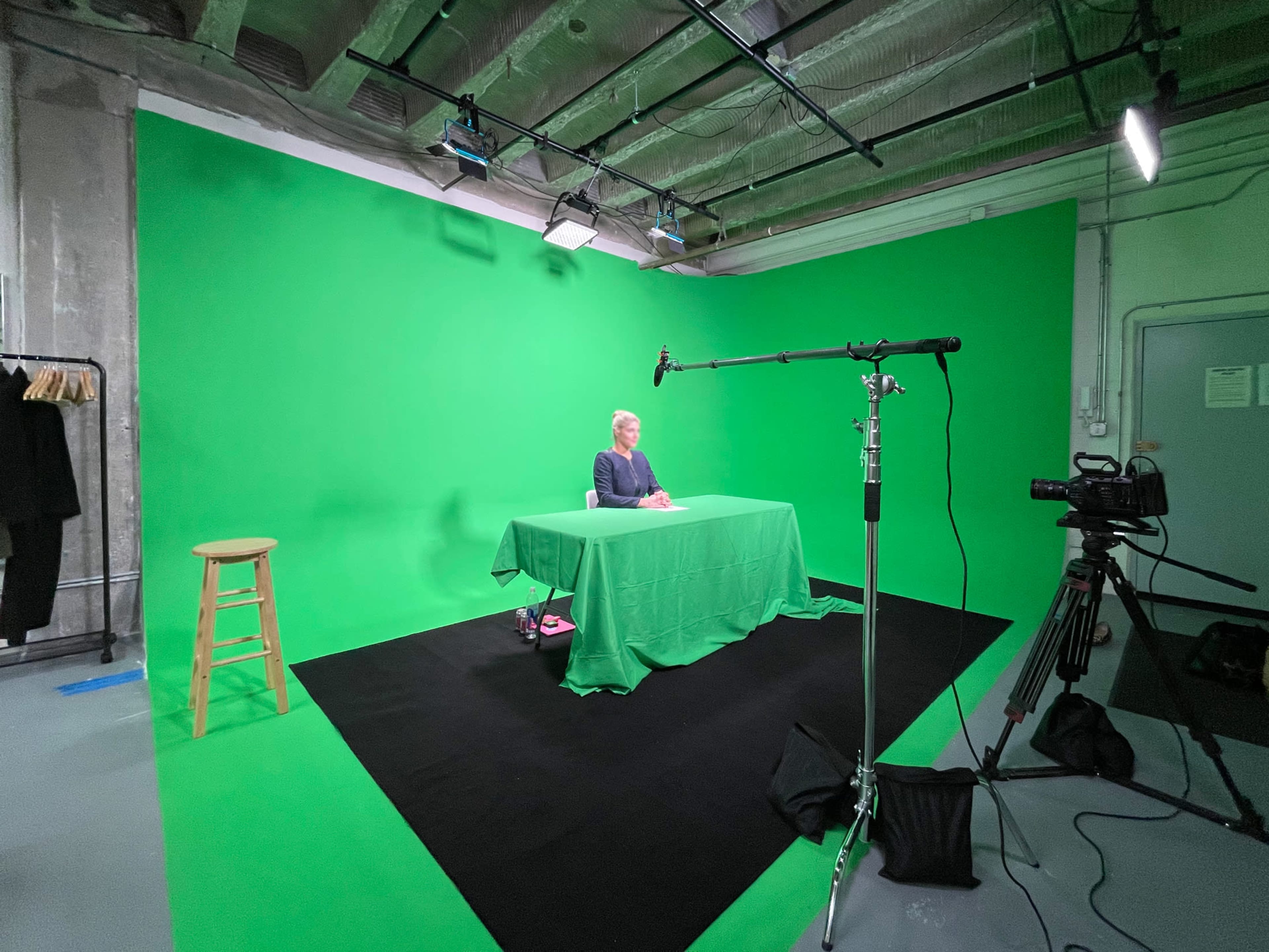A person sits at a table covered with a green cloth in a studio with a green backdrop and video equipment.
