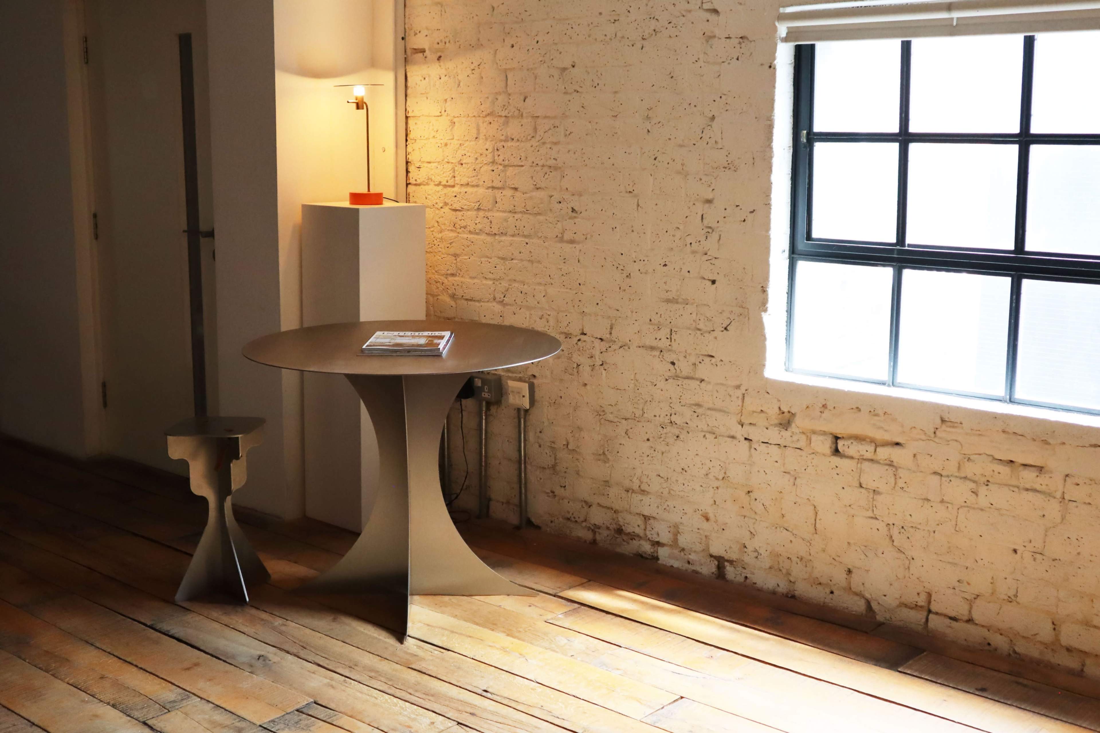 A round table and a slender stool are positioned near a large window on a wooden floor, with a lamp illuminating the space.