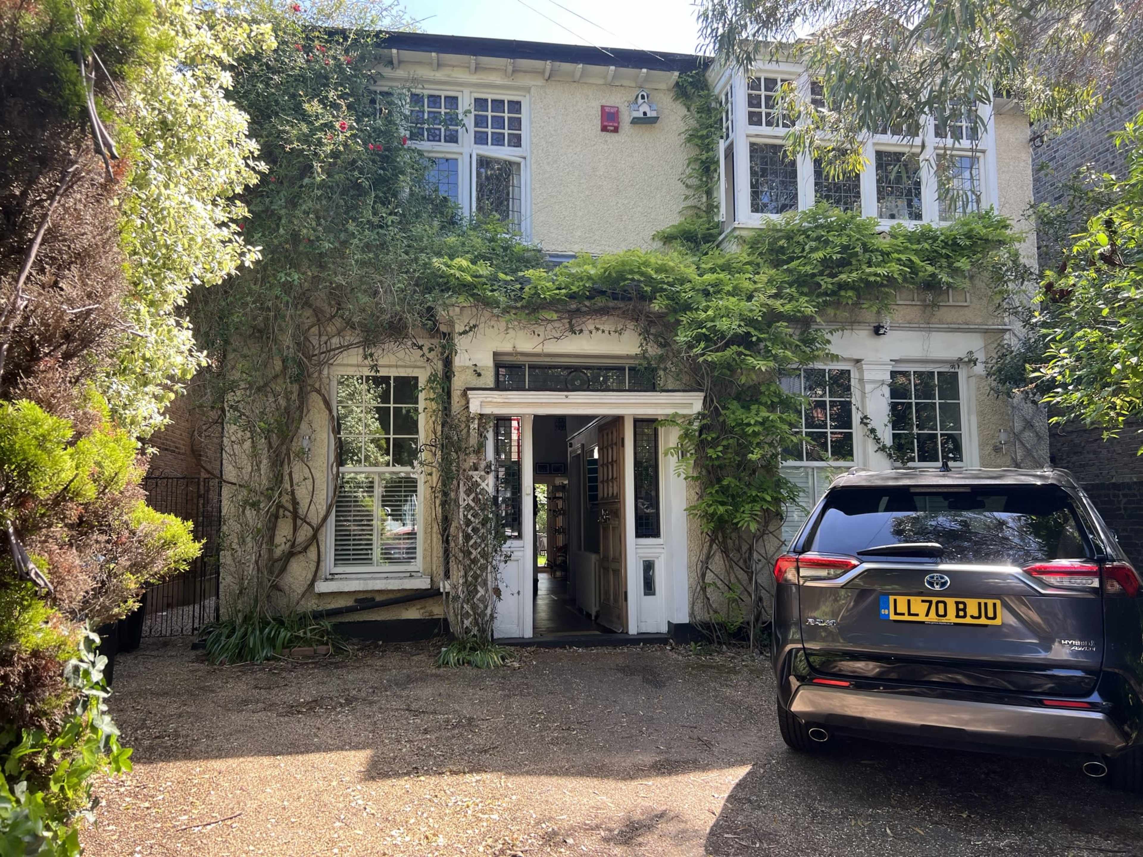 The image shows a two-story house covered in greenery, with a driveway leading up to the entrance and a parked black SUV nearby.