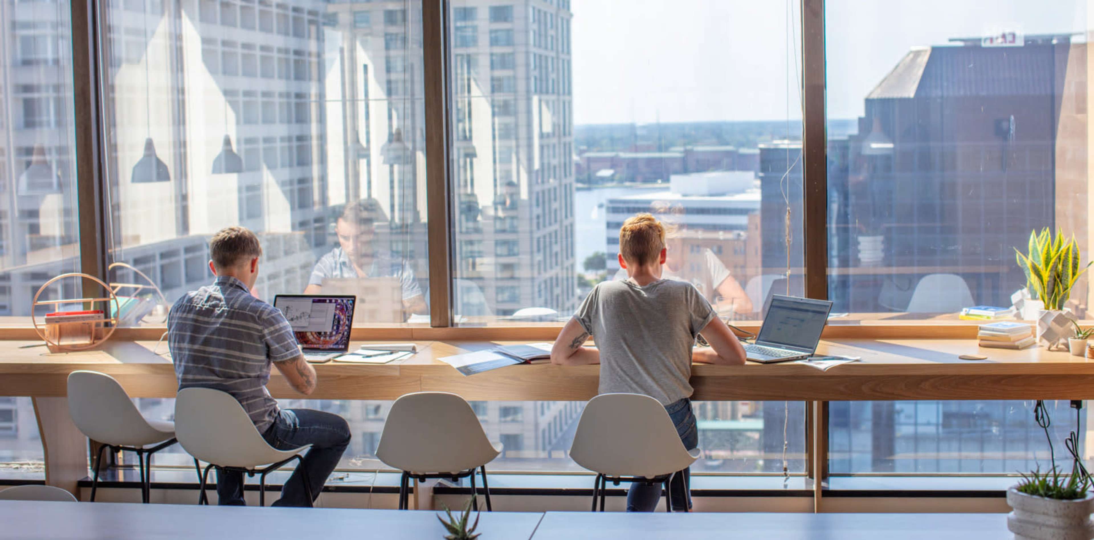Two men sit at a long wooden table by a large window, working on laptops with a city skyline visible outside.