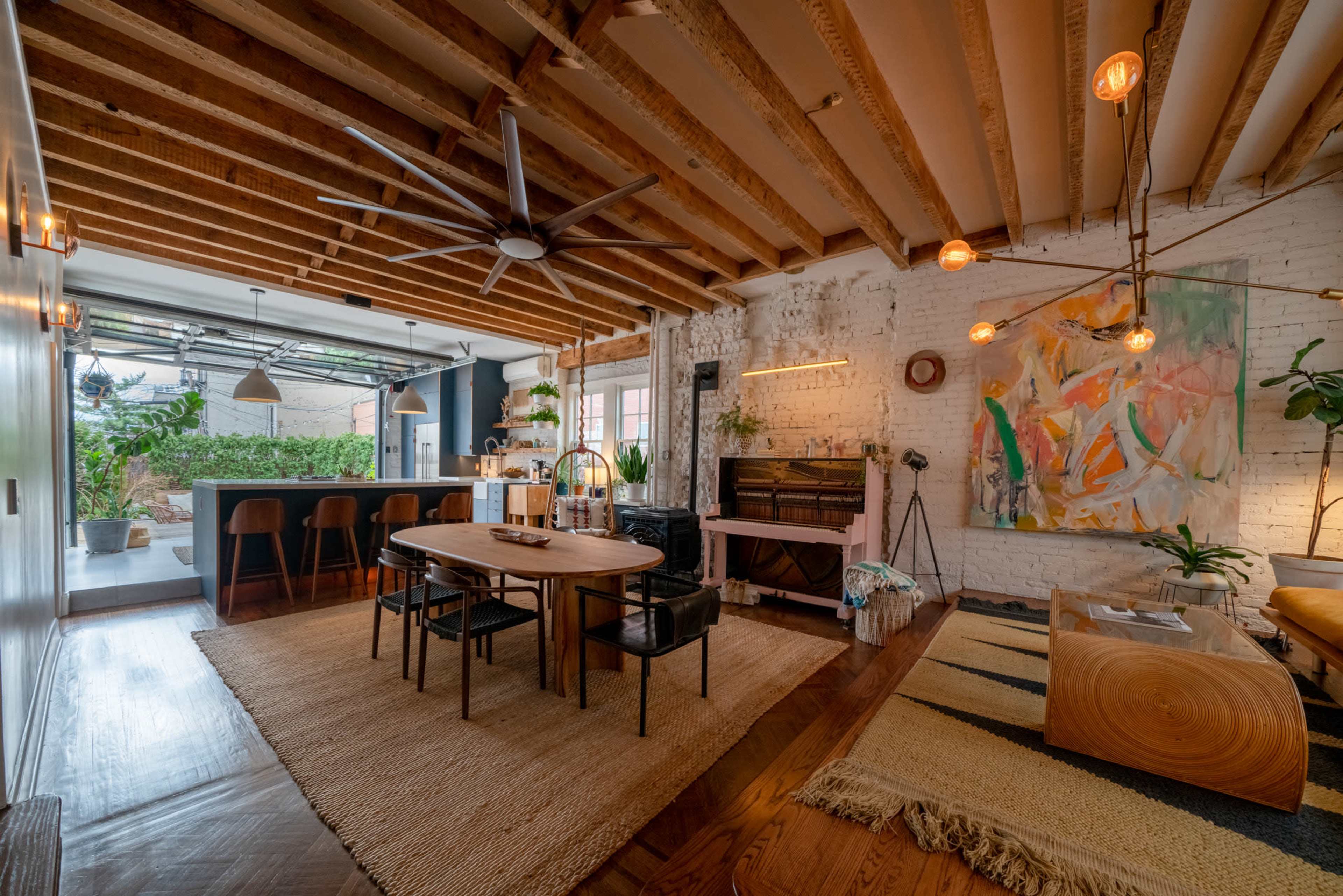 The scene features an open-concept living area with a kitchen, dining table, and vibrant artwork, highlighted by exposed wooden beams and ample natural light.