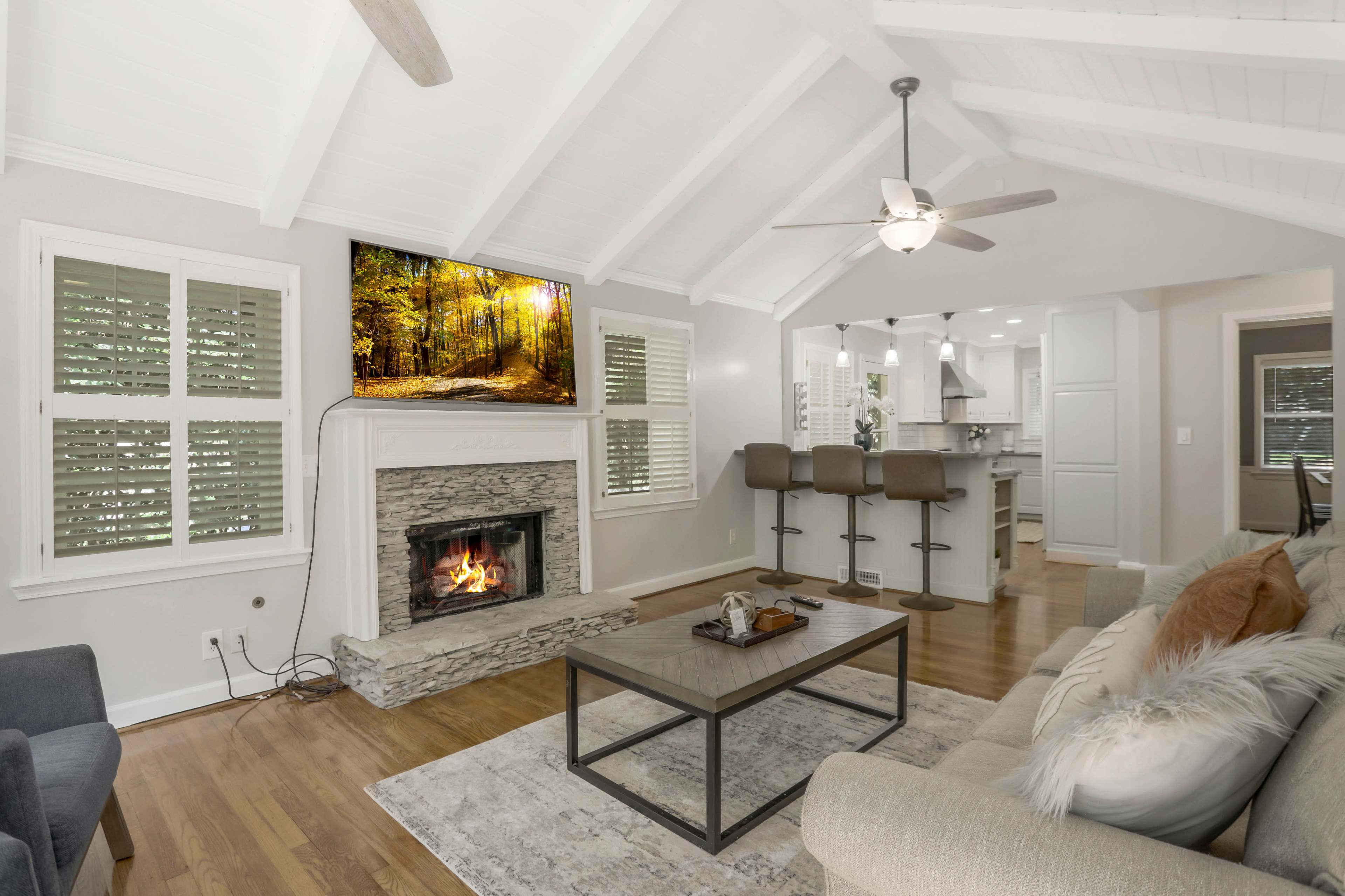 A modern living room features a stone fireplace, a flat-screen TV mounted above it, and an open layout that leads to a kitchen with bar seating.