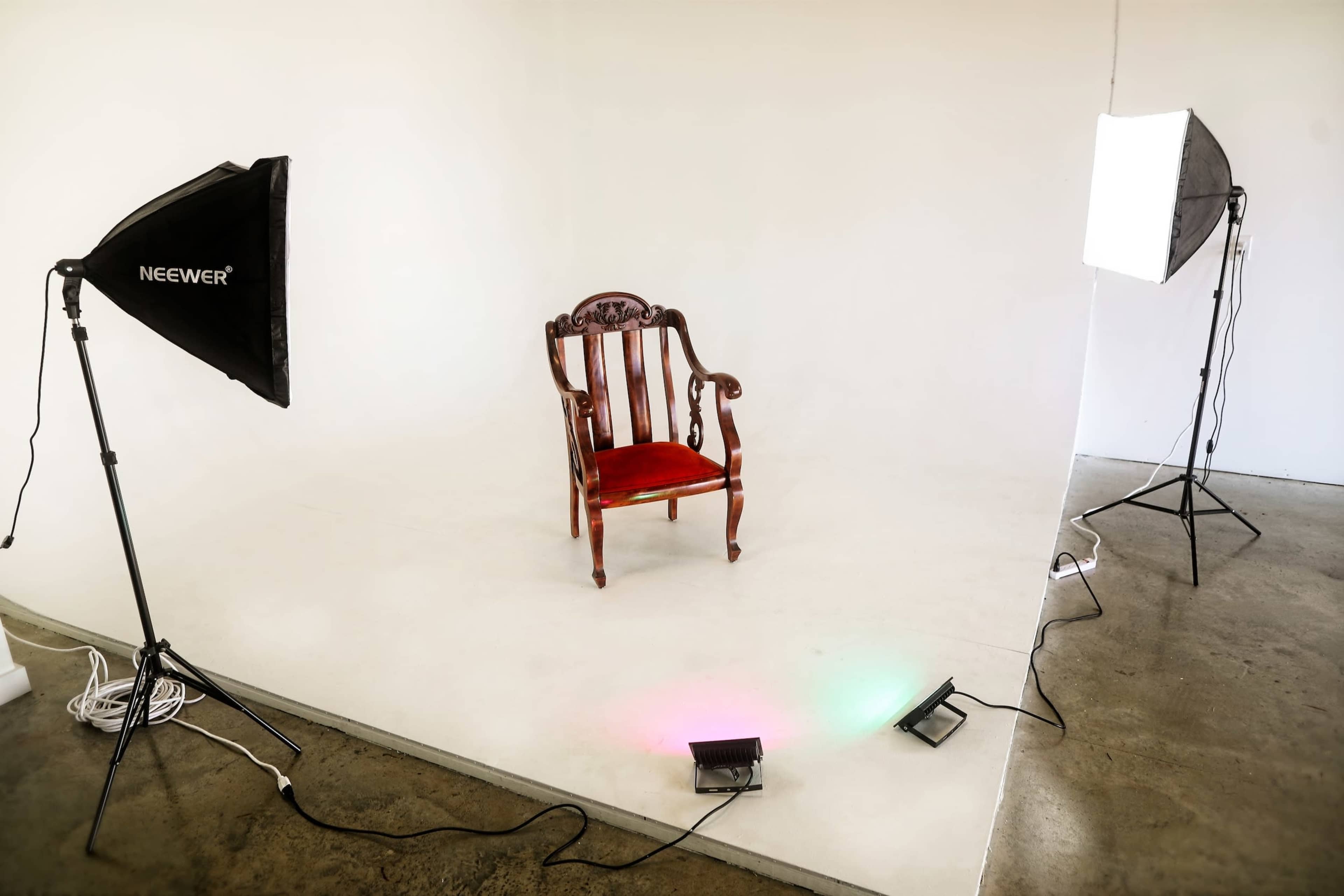 A wooden chair with red upholstery positioned on a plain, light-colored floor, surrounded by softbox lighting setups on either side.