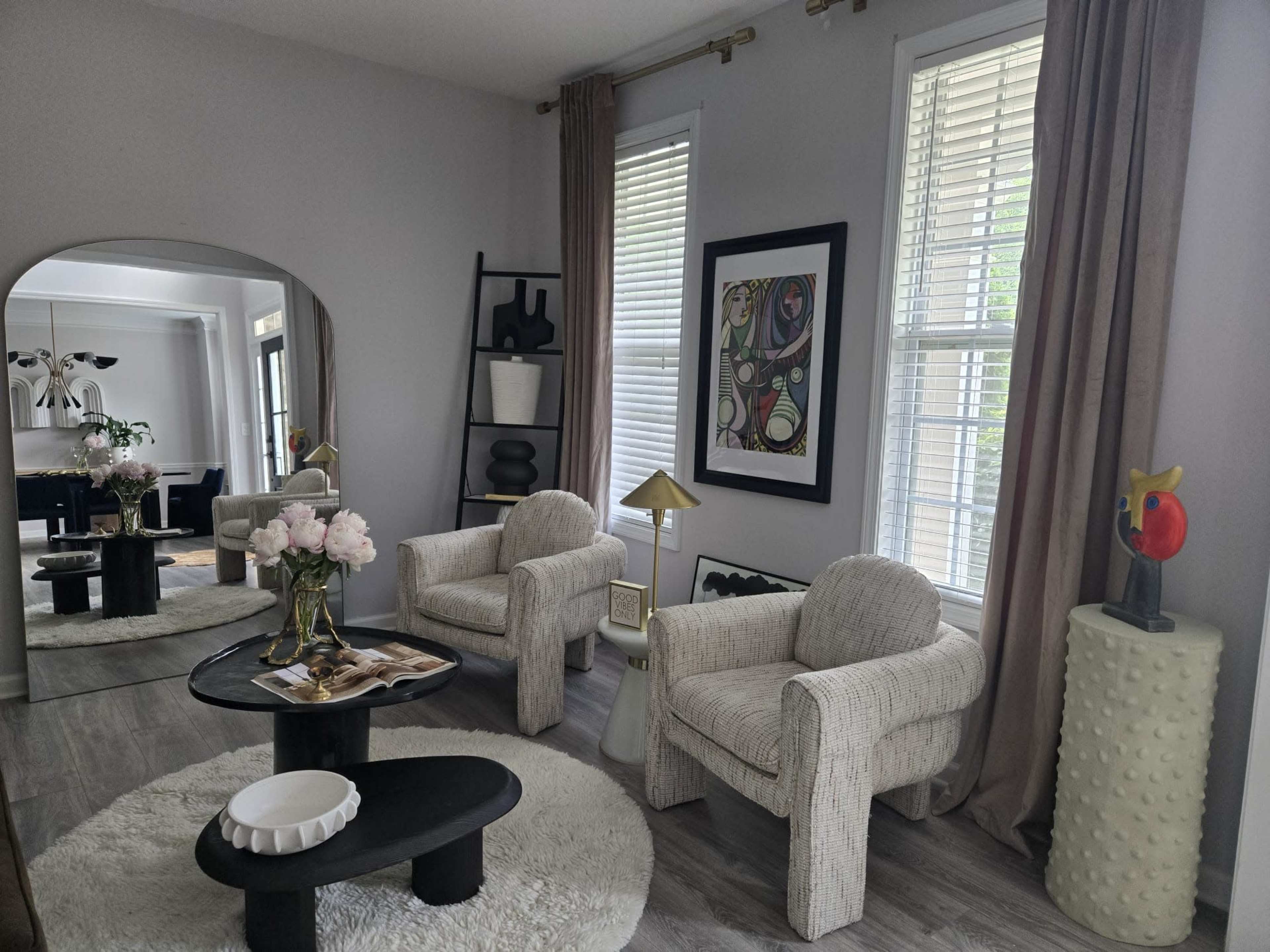 The image shows a modern living room with two upholstered chairs, a round coffee table, and art pieces on the walls, all set against large windows that allow natural light to enter.