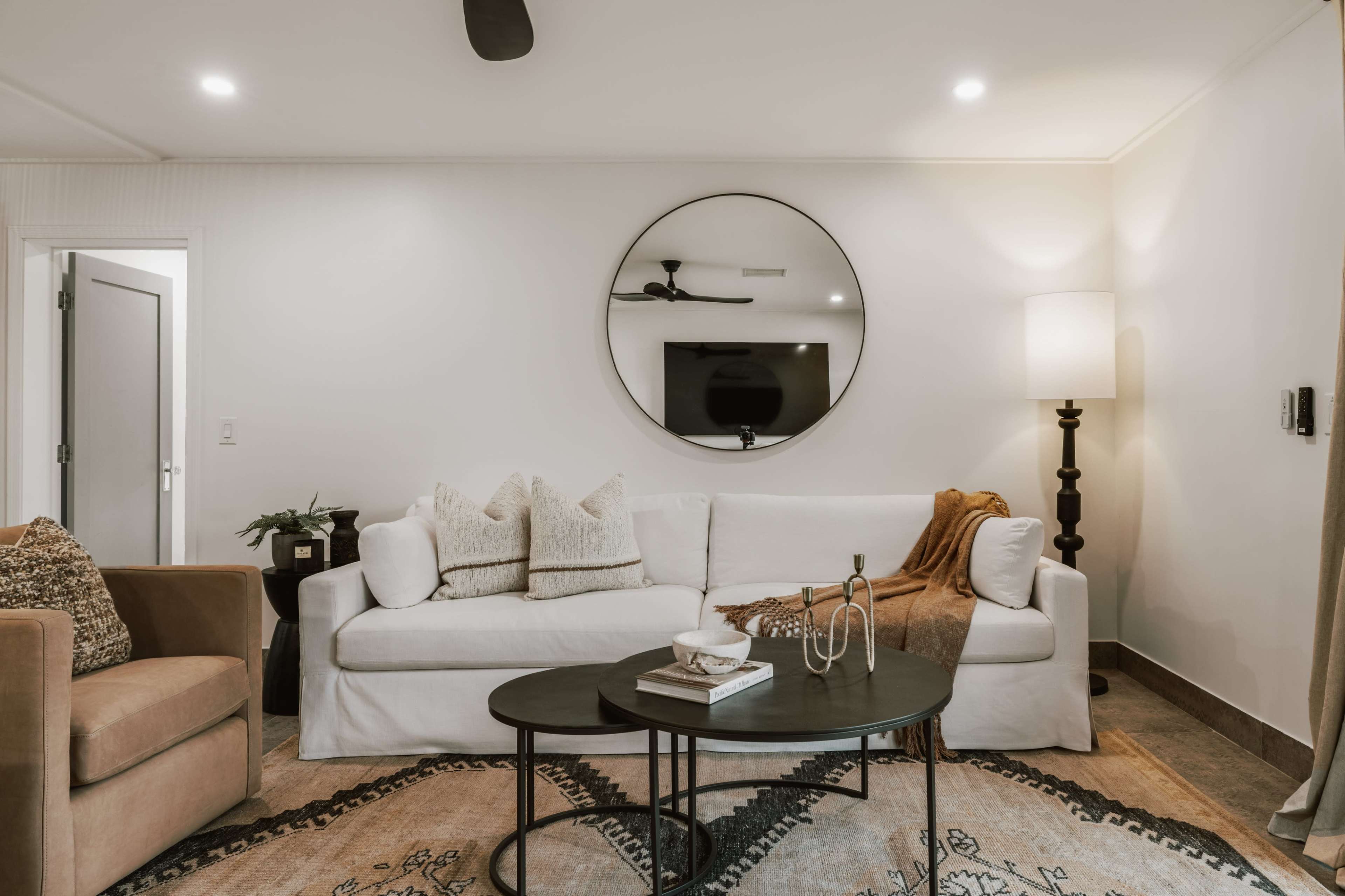 The scene features a modern living room with a white sofa, round mirror, dark coffee table, and a stylish floor lamp beside a wall-mounted television.