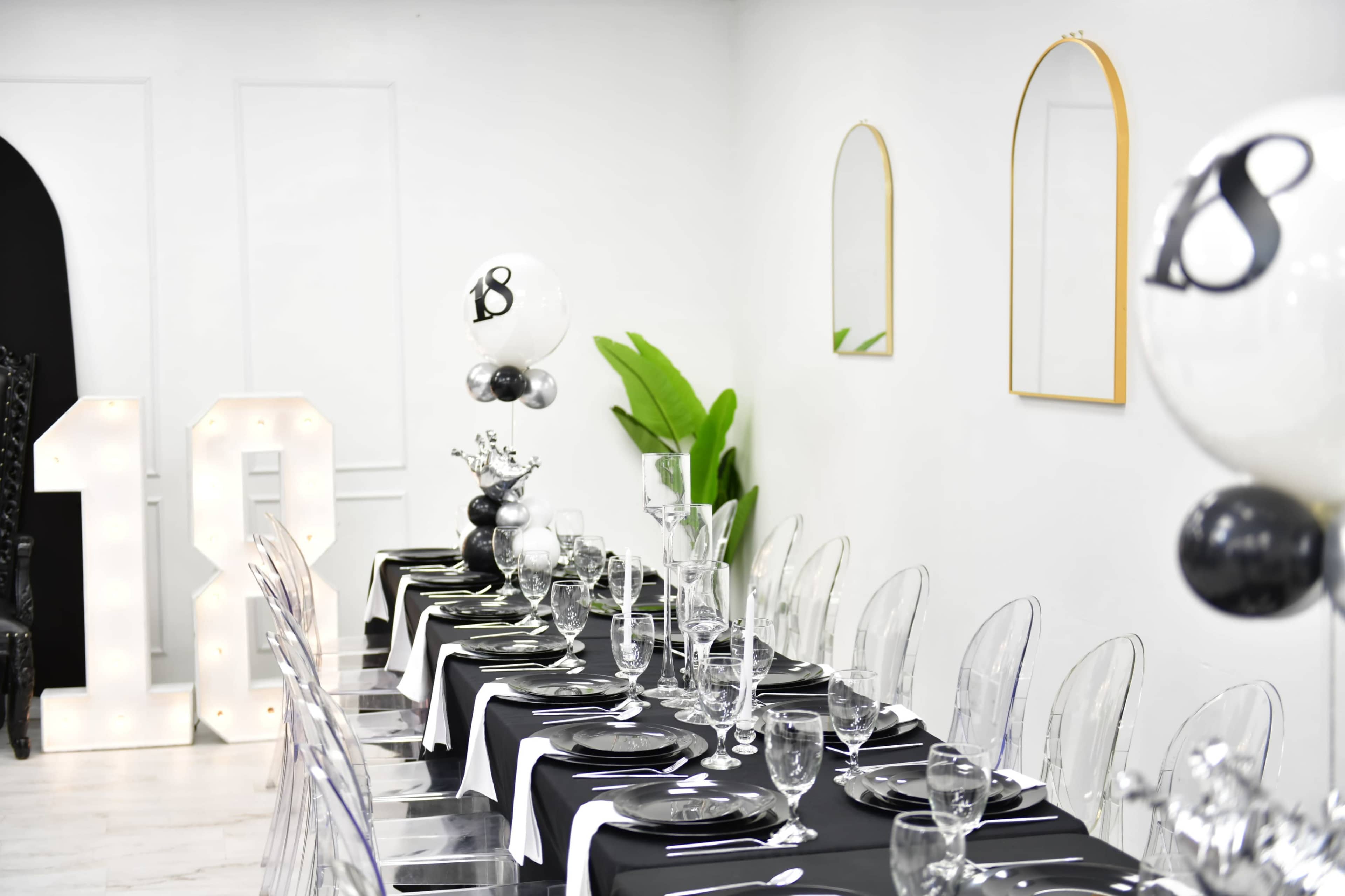 The image shows a decorated dining area with a long, black and white table set with glassware and elegant tableware, flanked by clear acrylic chairs and adorned with balloons featuring the number "18."