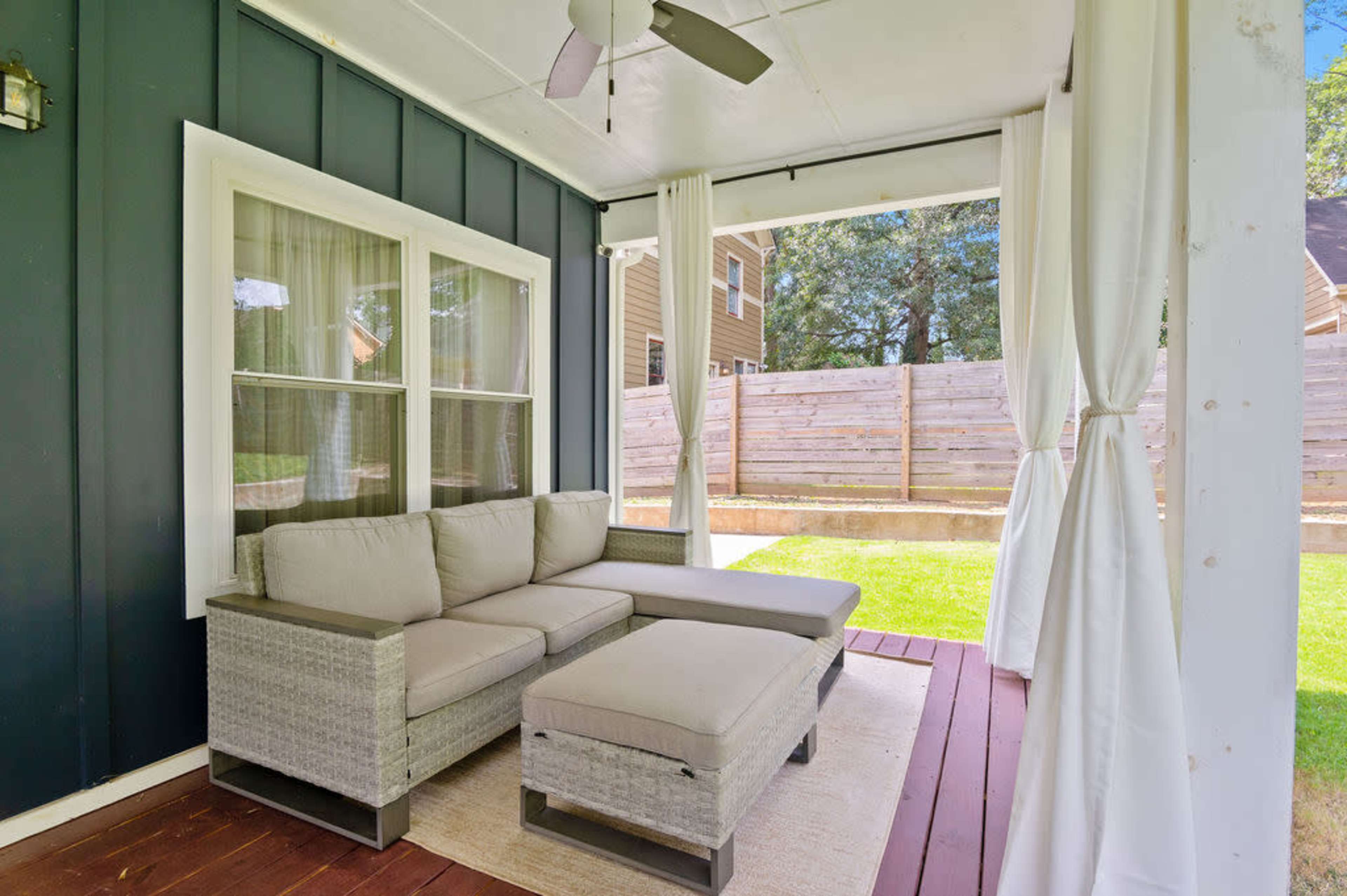 A covered porch features a gray sectional sofa and a matching ottoman, with sheer curtains flanking the windows.