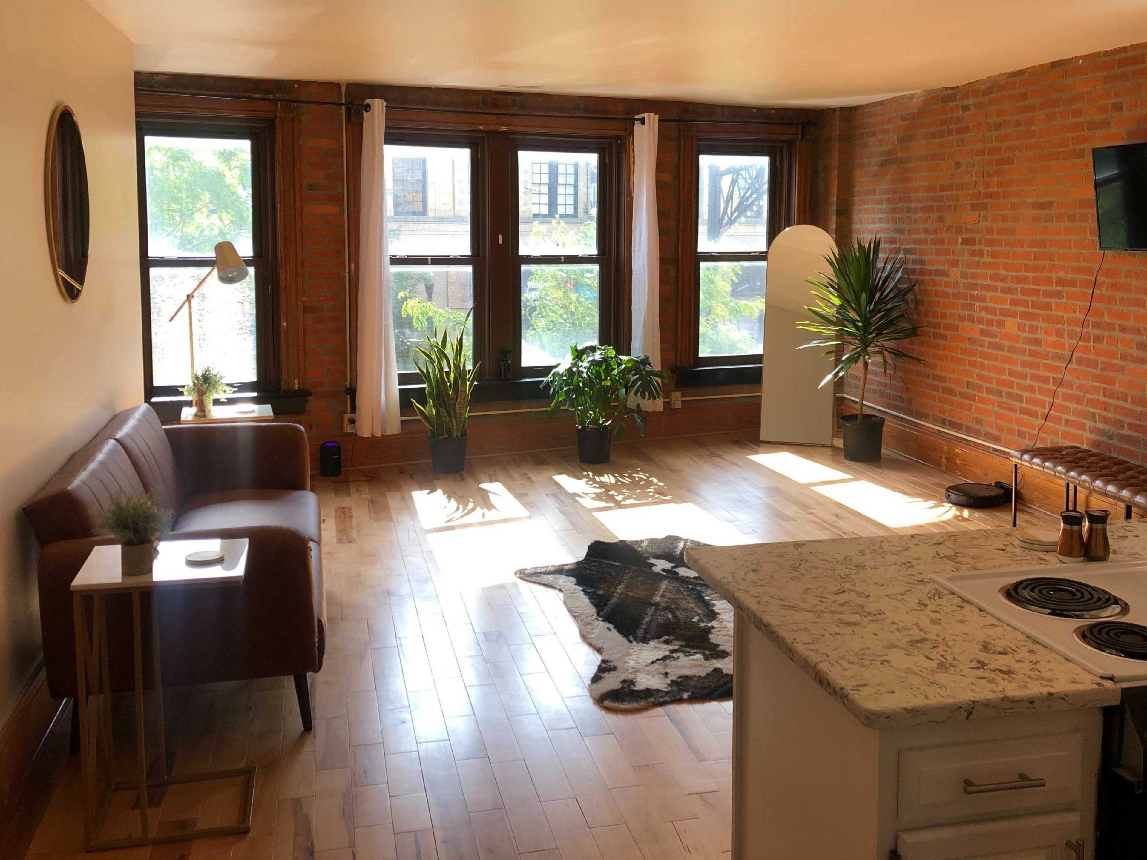 A well-lit living room with exposed brick walls, a brown leather couch, potted plants, a small side table, and a kitchen area.