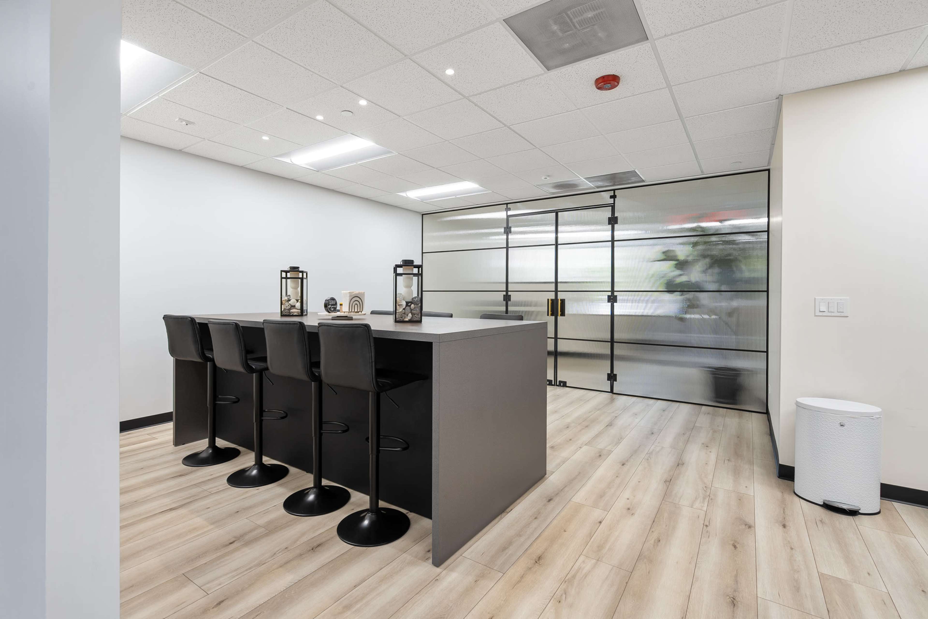 The image shows a modern office space with a bar-style area featuring four black stools and a partitioned glass wall in the background.