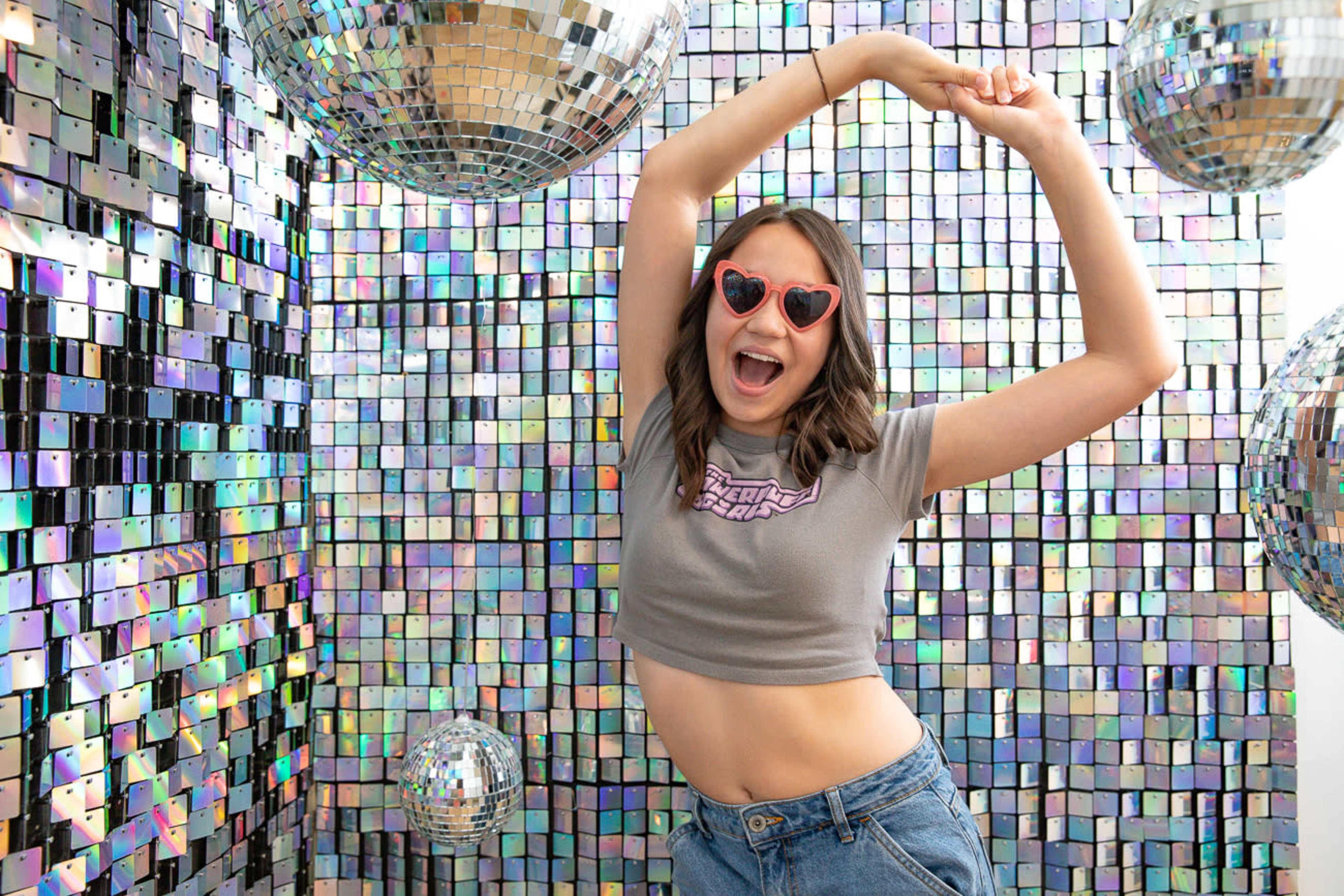 A woman in a cropped shirt dances playfully in front of a shimmering, reflective wall adorned with disco balls.