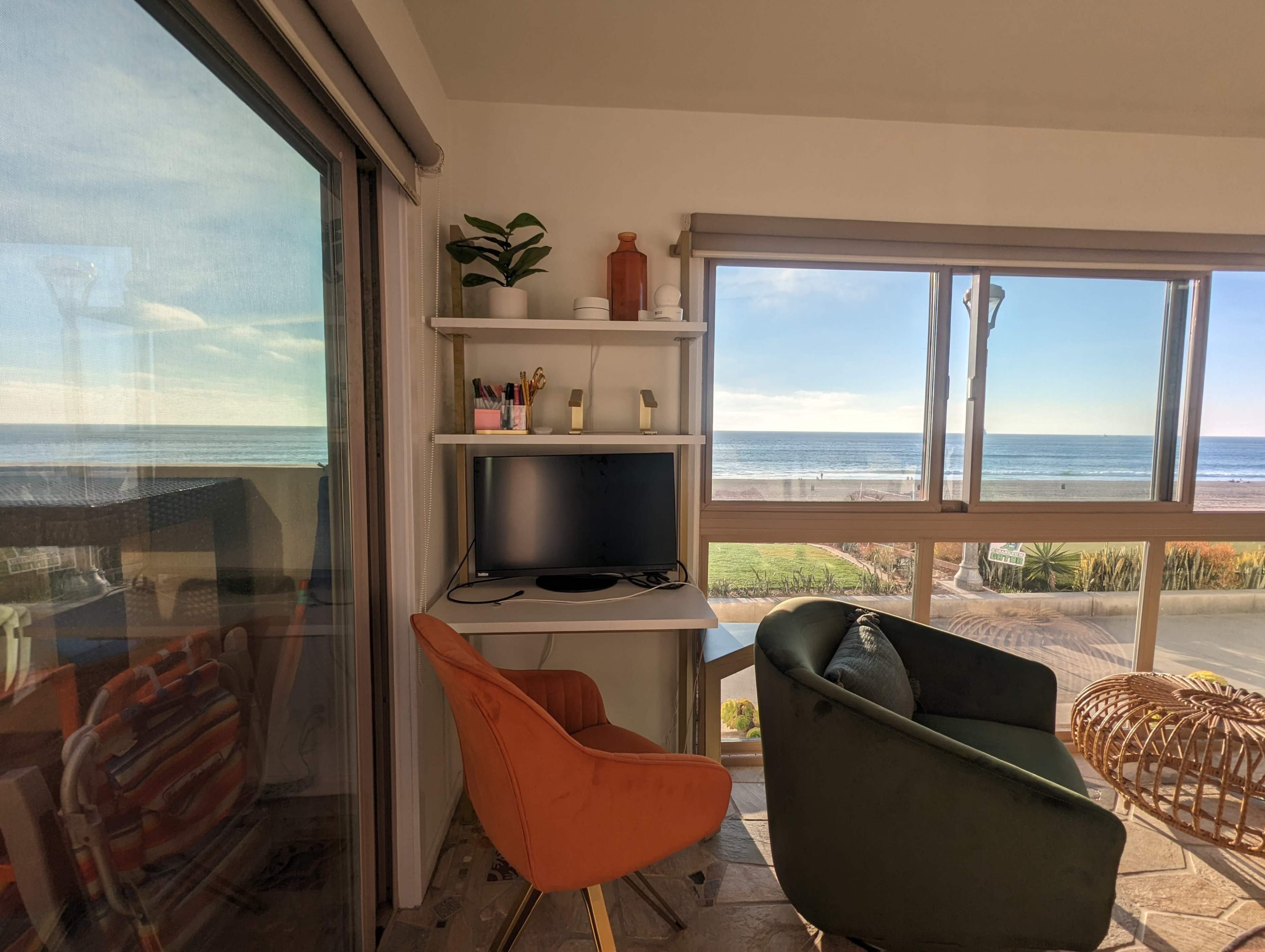 A desk with a computer screen and an orange chair is positioned by a large window overlooking the ocean.