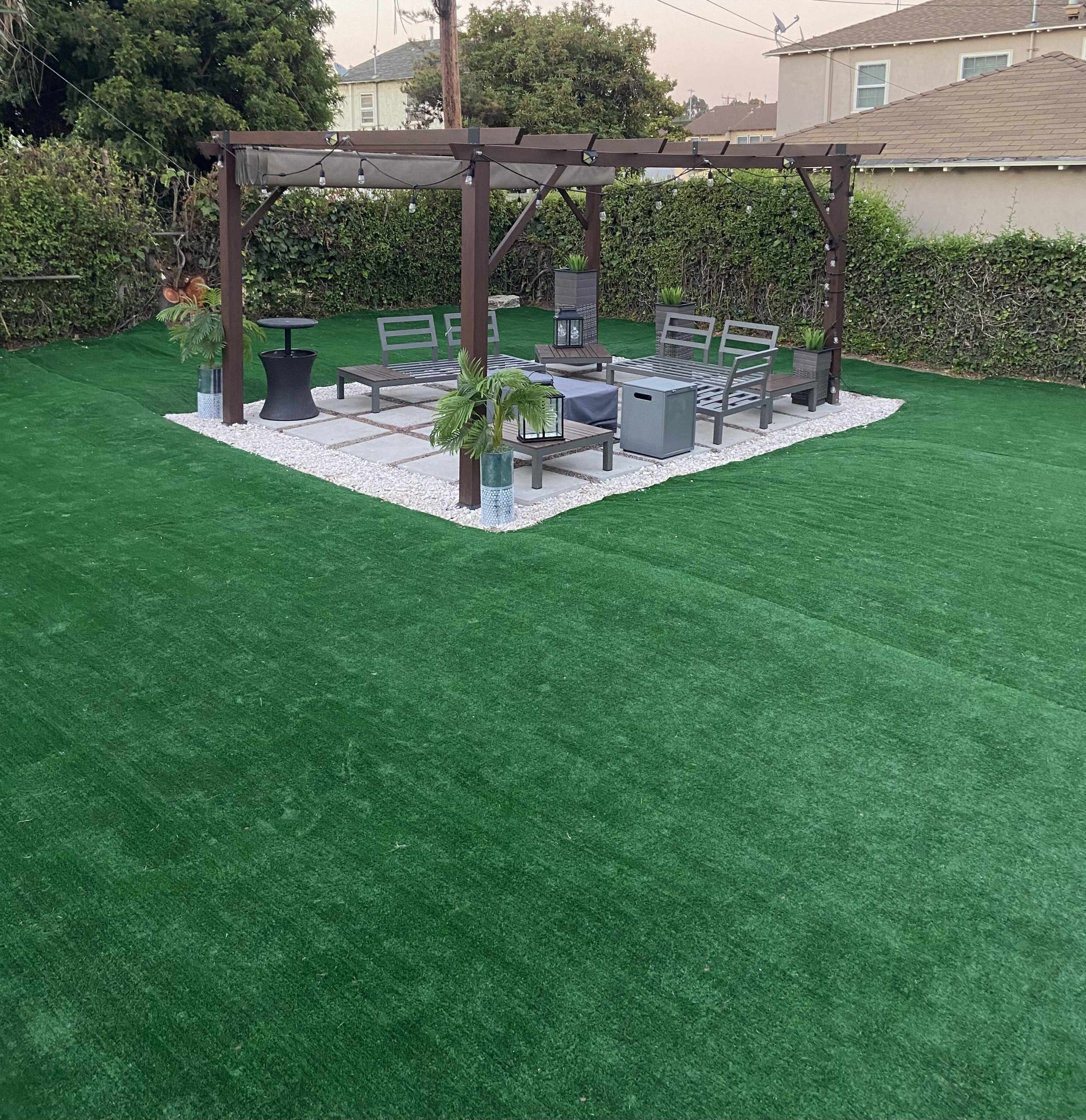 The image shows a yard with artificial grass featuring a shaded seating area under a pergola, surrounded by greenery.