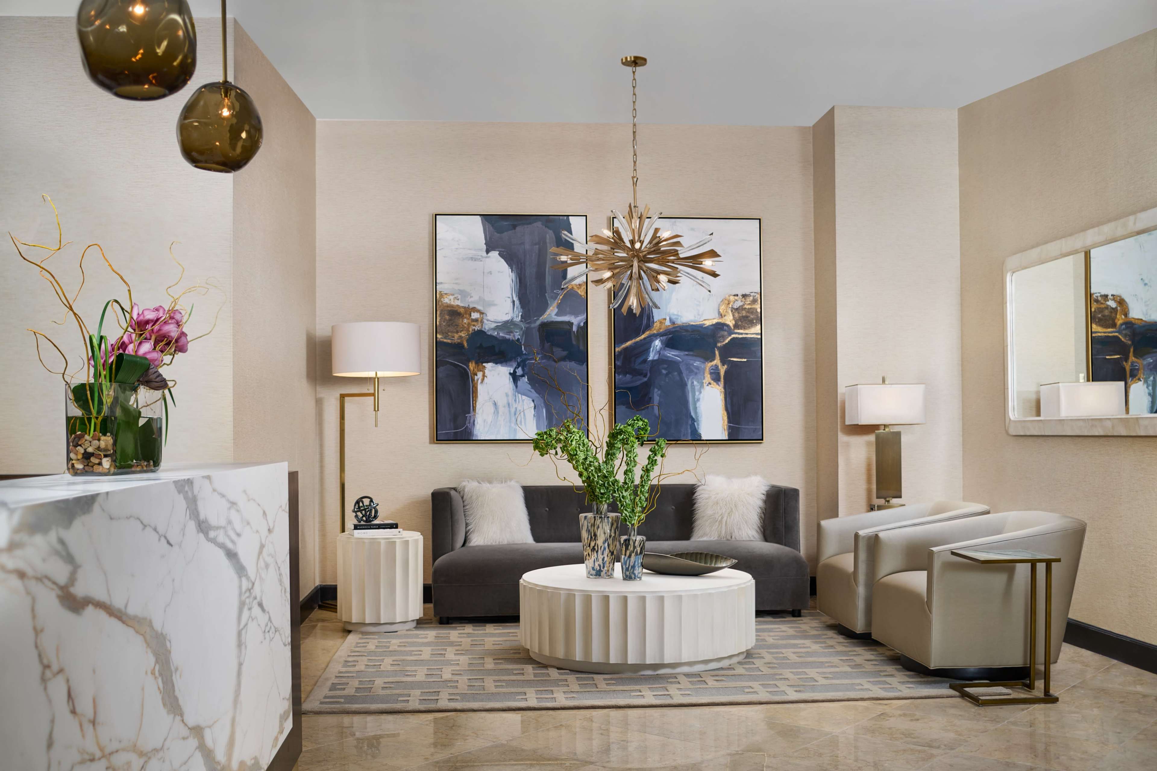 A modern lobby area featuring a gray sofa, two armchairs, a round coffee table, decorative artwork on the walls, and a marble reception desk with plants and lighting accents.