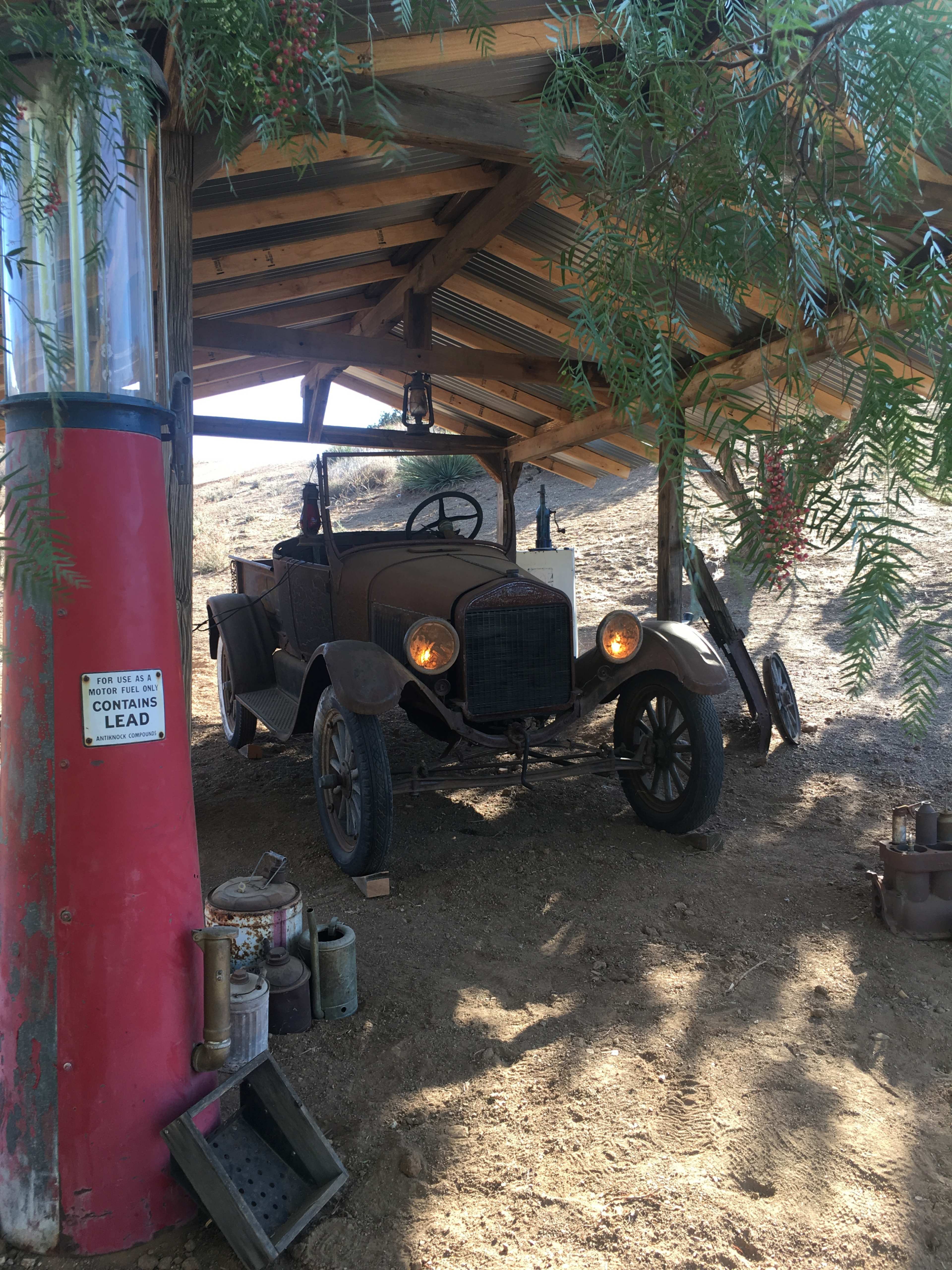 An old, weathered car is parked under a wooden shelter next to a vintage gas pump surrounded by dirt and sparse vegetation.