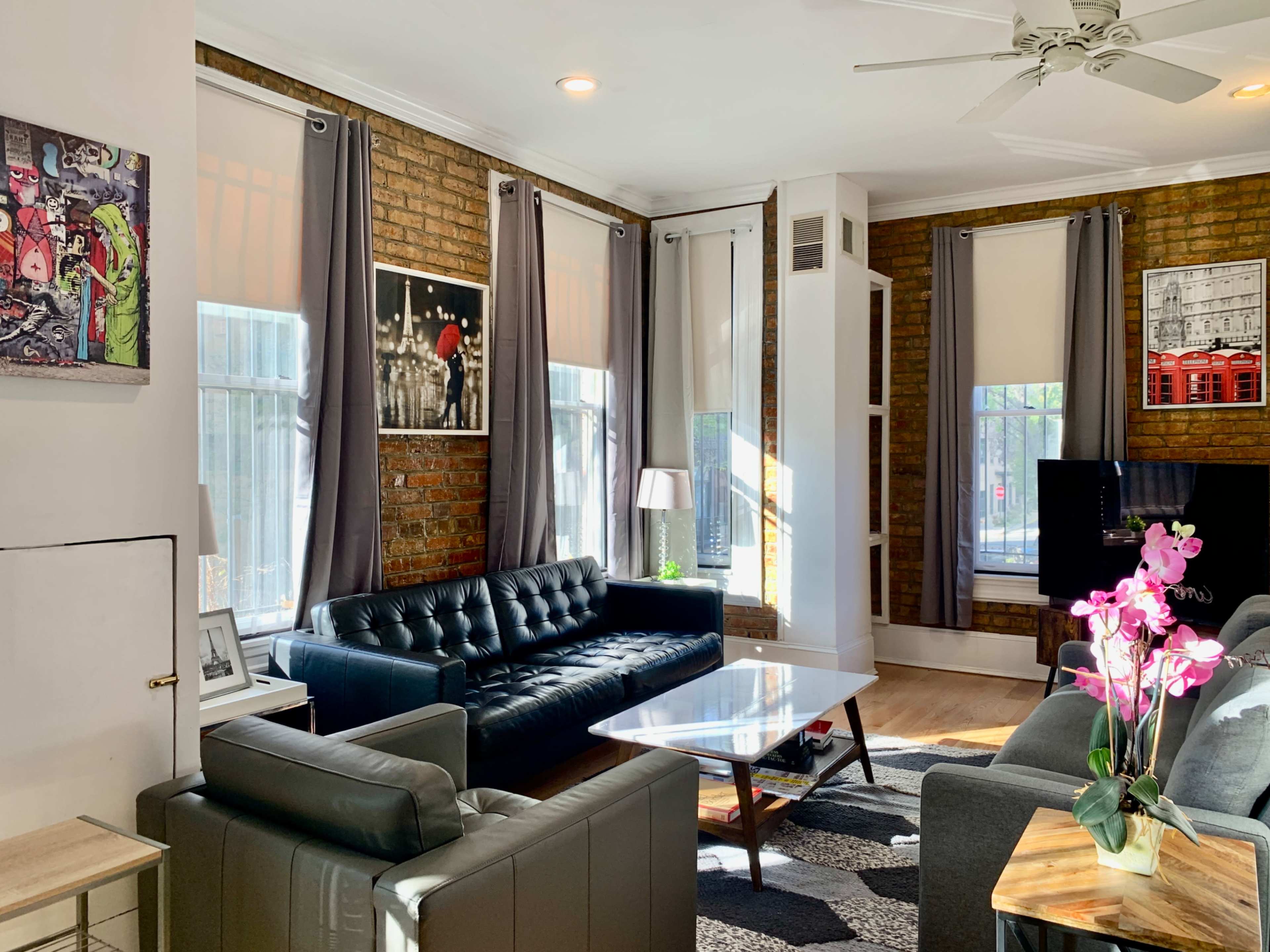 A bright living room features two sofas, a glass coffee table, and large windows with light filtering through sheer curtains.