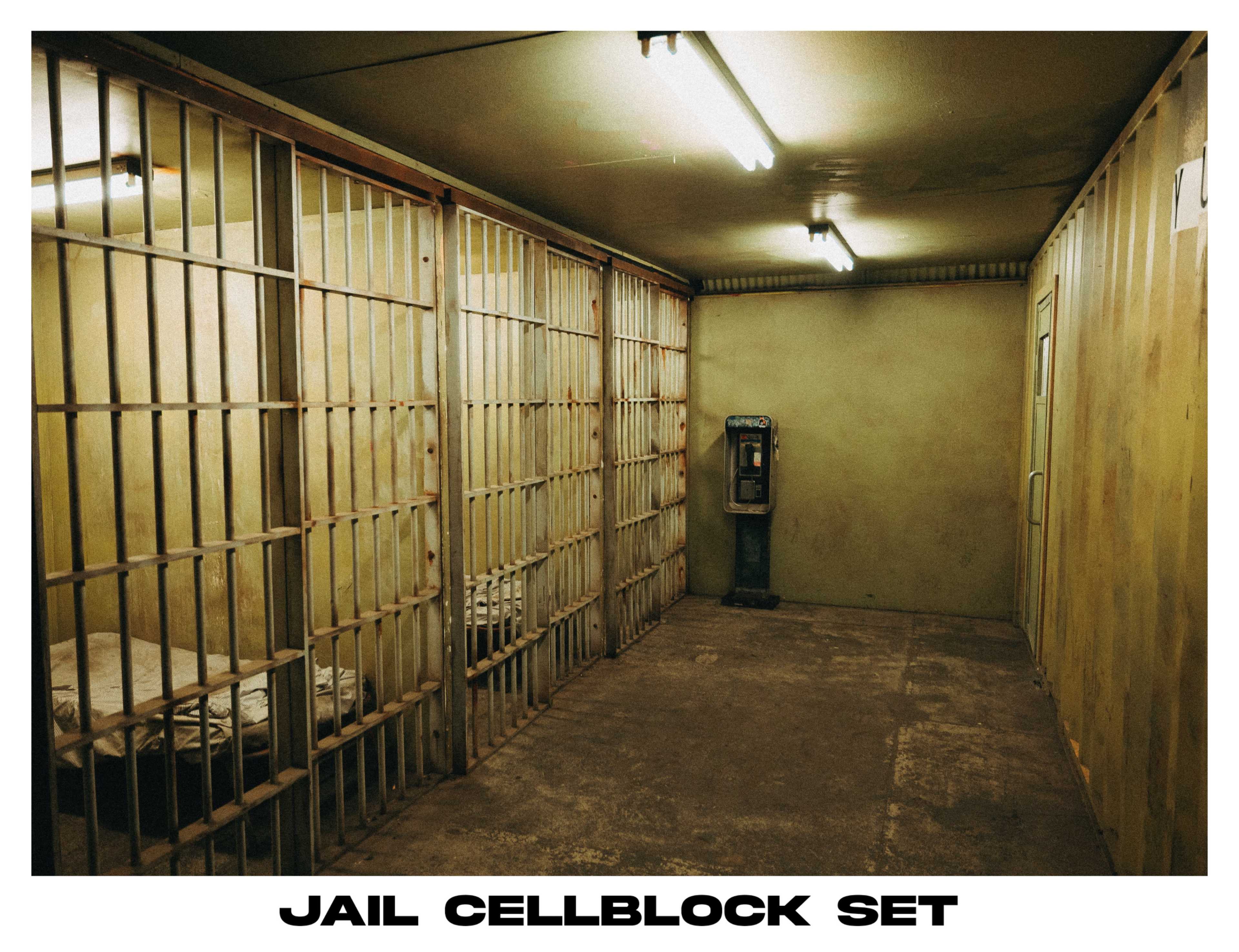 The image depicts a jail cellblock set featuring metal bars, a bed inside a cell, and a payphone against a wall.