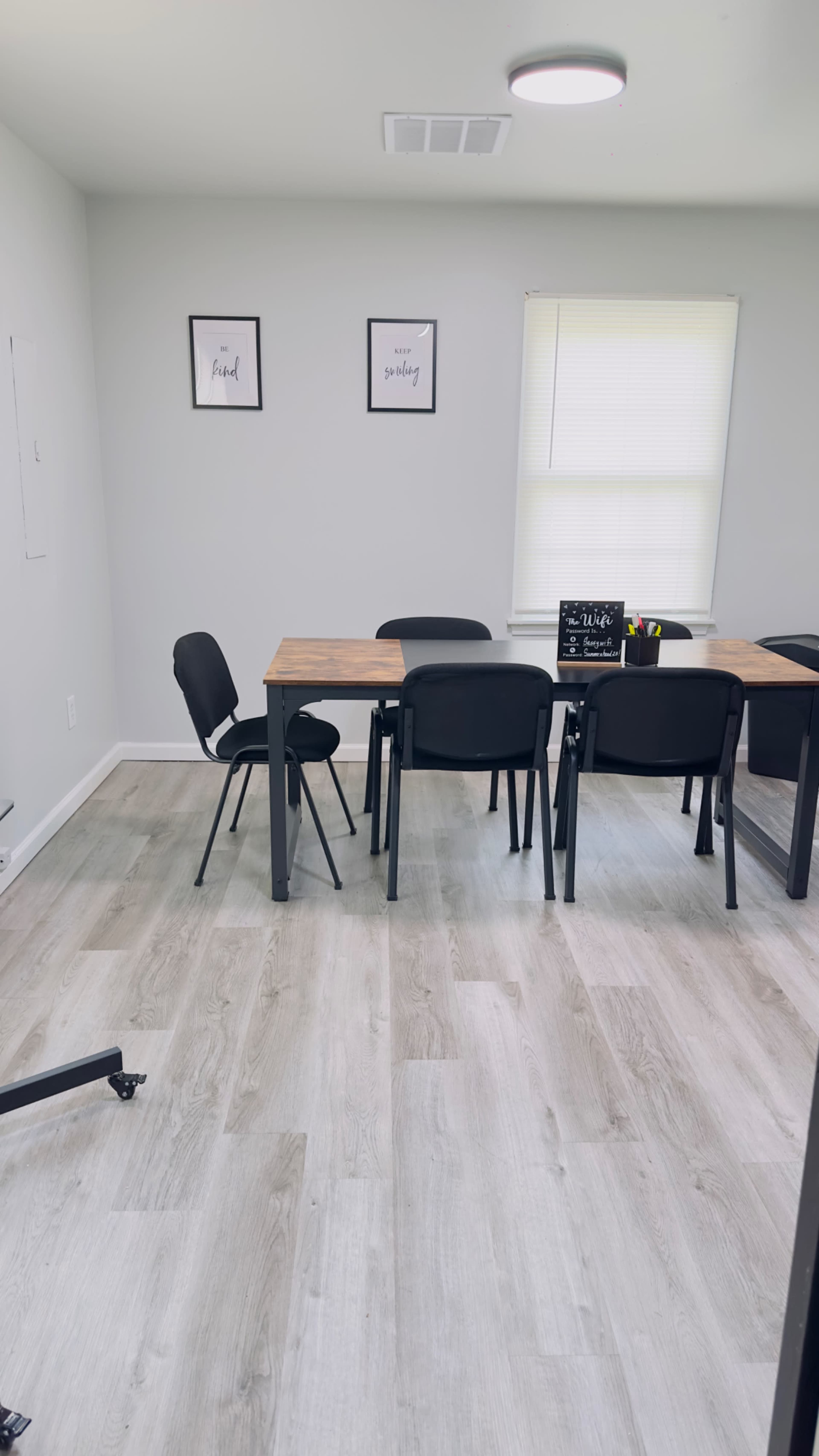 A light-colored room with a wooden table surrounded by black chairs and two framed prints on the wall.