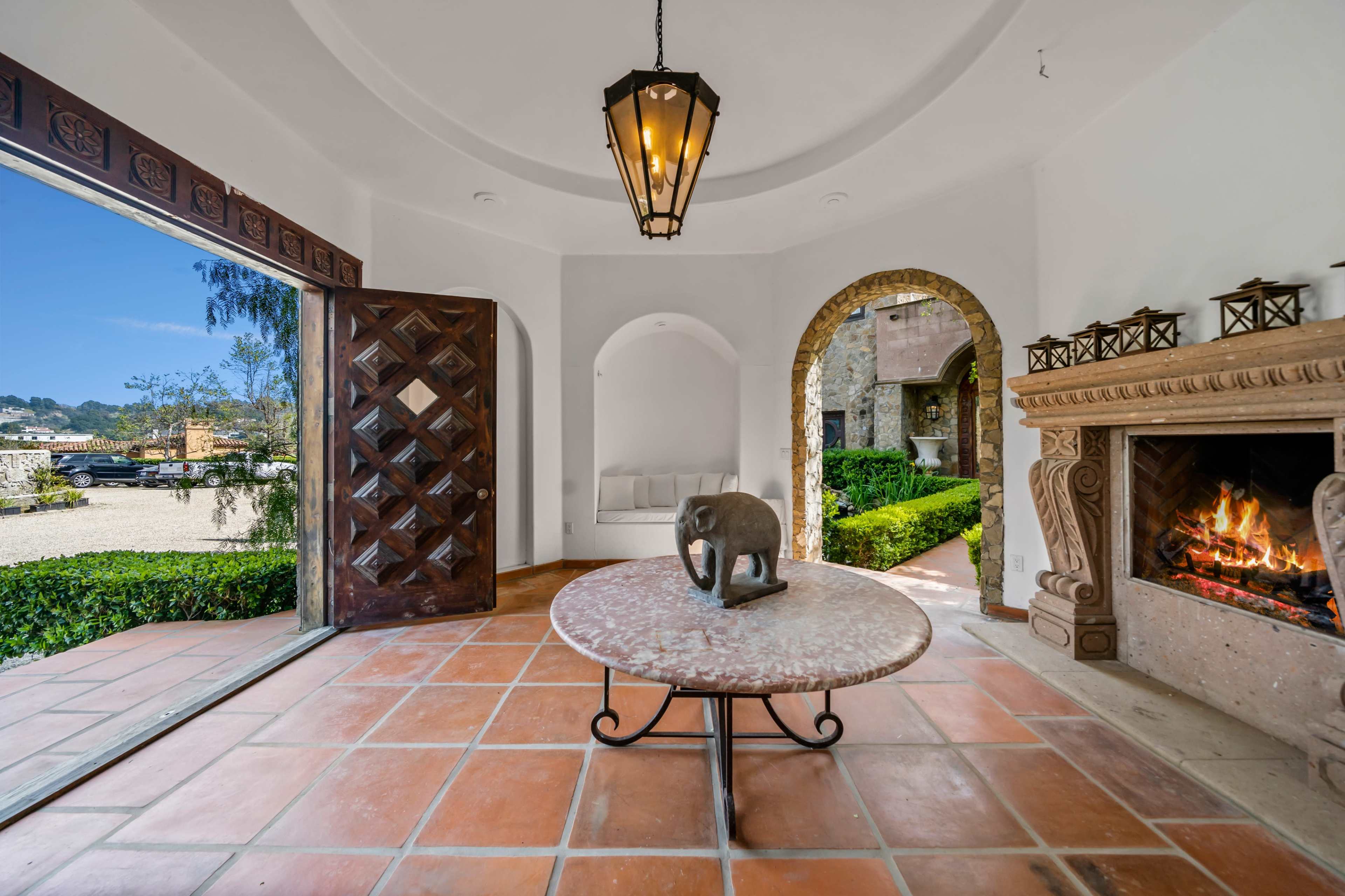 The image shows a spacious entryway featuring a round table with an elephant sculpture, a stone fireplace, and large doors that open to a landscaped outdoor area.