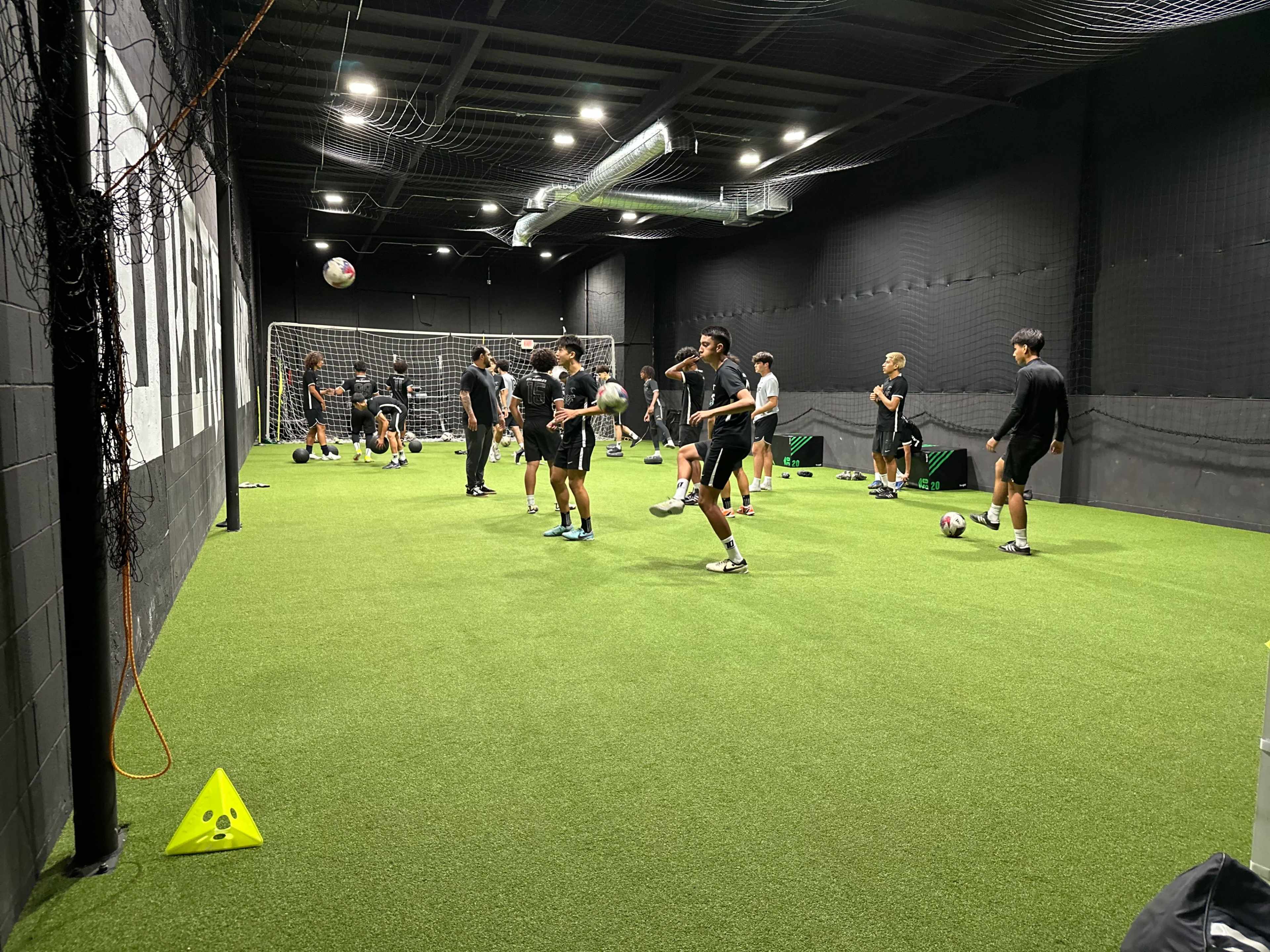 A group of athletes is practicing soccer drills in a carpeted indoor training facility.