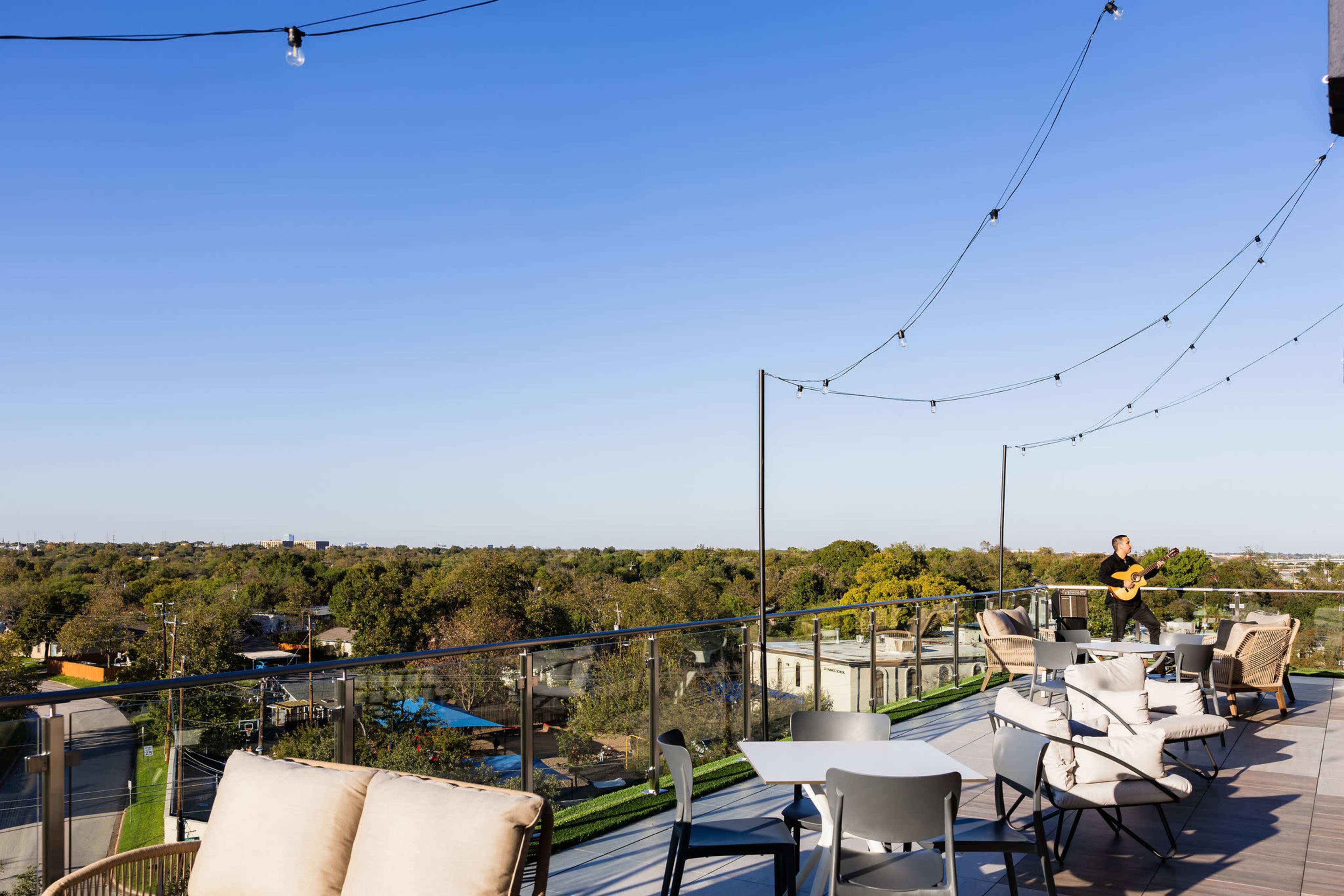 A rooftop terrace features comfortable seating and string lights, overlooking a landscape of trees and buildings under a clear blue sky.