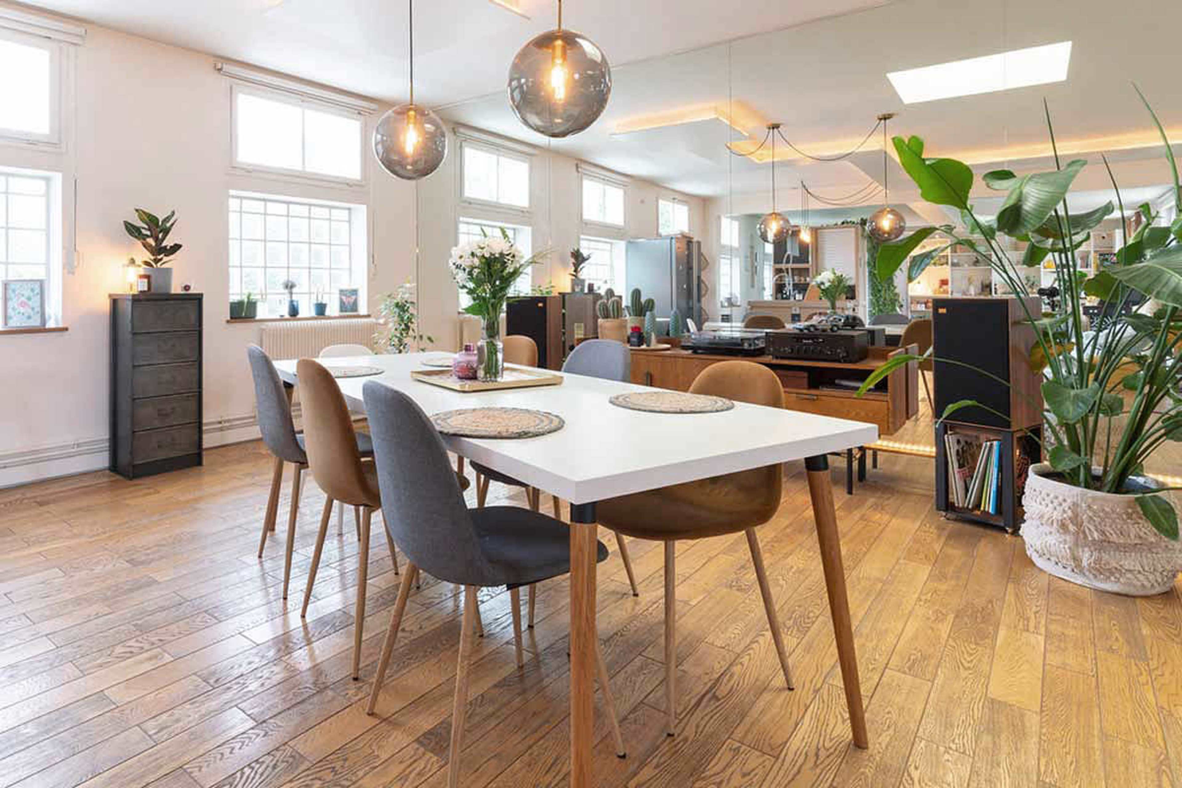 The image shows a modern dining area with a white table surrounded by chairs, large windows, and indoor plants, all set in a spacious, well-lit room with wooden flooring.