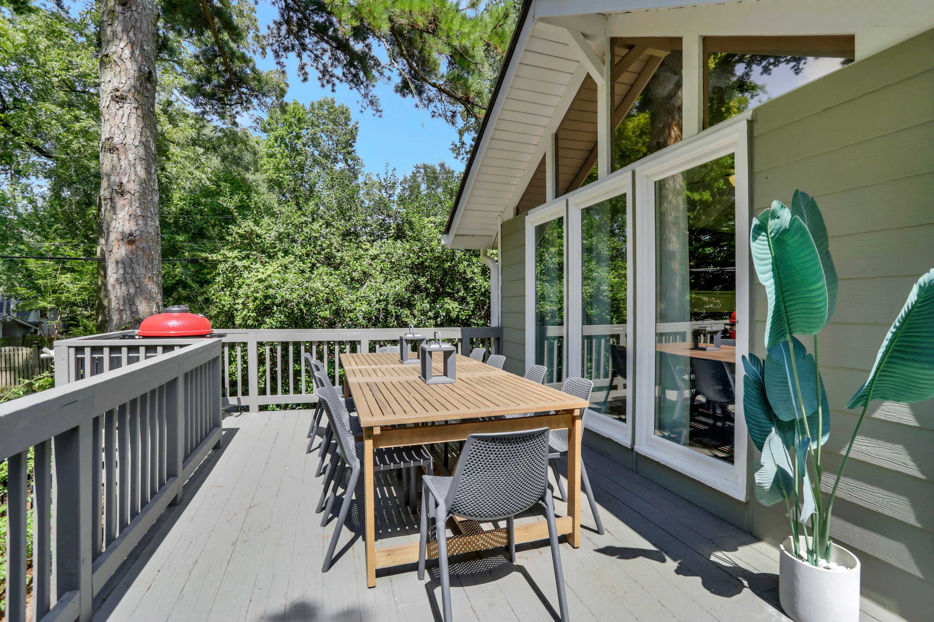 A wooden deck features a table with six chairs, a lantern, and a potted plant, surrounded by trees and a red grill in the background.