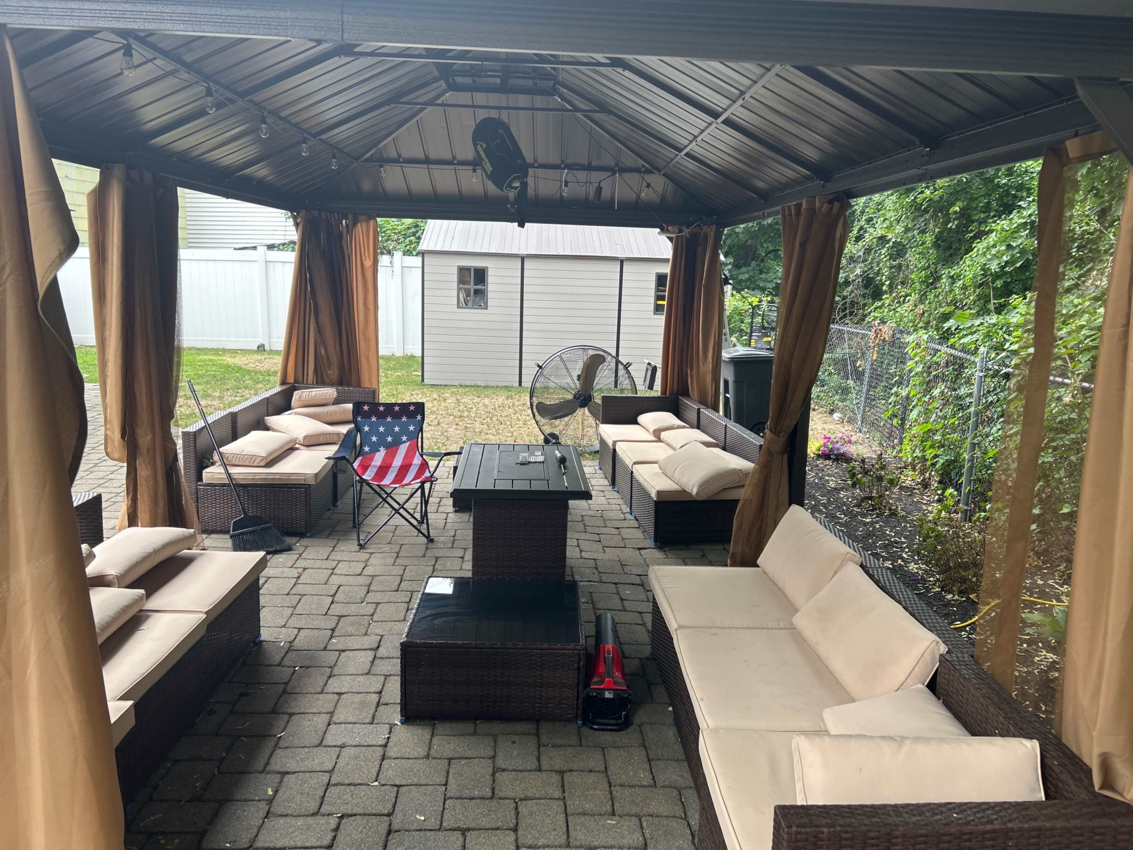 The image shows a covered outdoor seating area with a rectangular table, cushioned chairs, and a fan, surrounded by a gravel flooring and a fence.