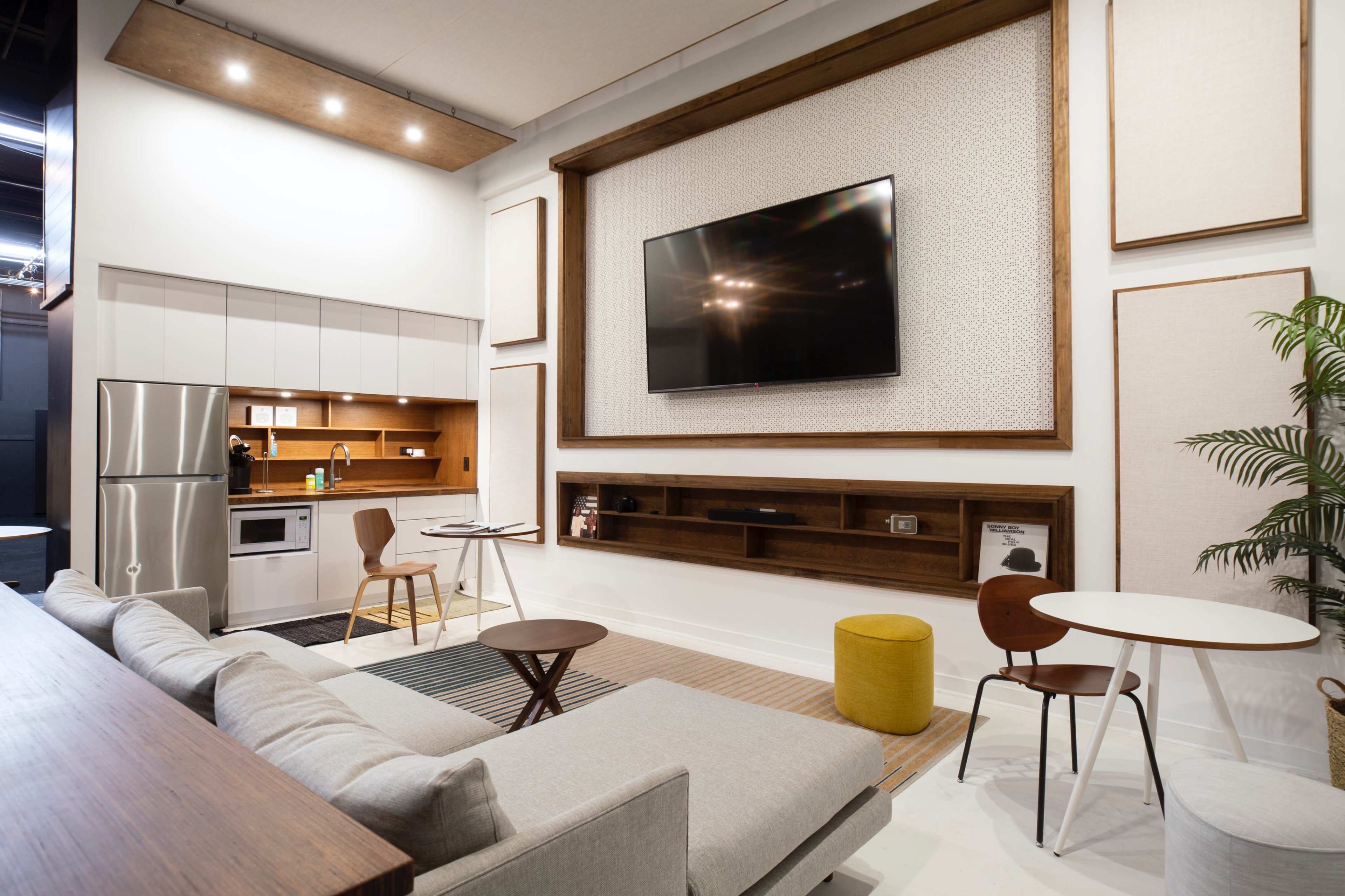 The image shows a modern living space featuring a kitchen area with stainless steel appliances, a wall-mounted television, and a seating area with a sectional sofa and circular dining table.