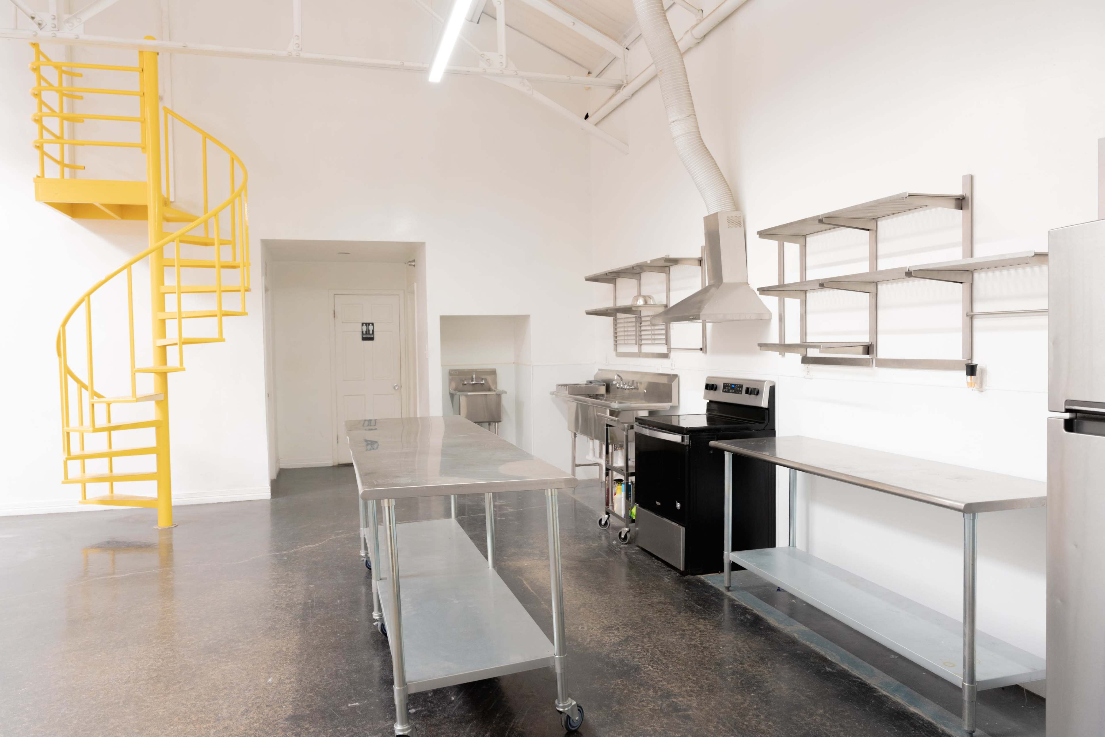 The image depicts a modern kitchen with stainless steel counters, a yellow spiral staircase, and an overhead ventilation system.