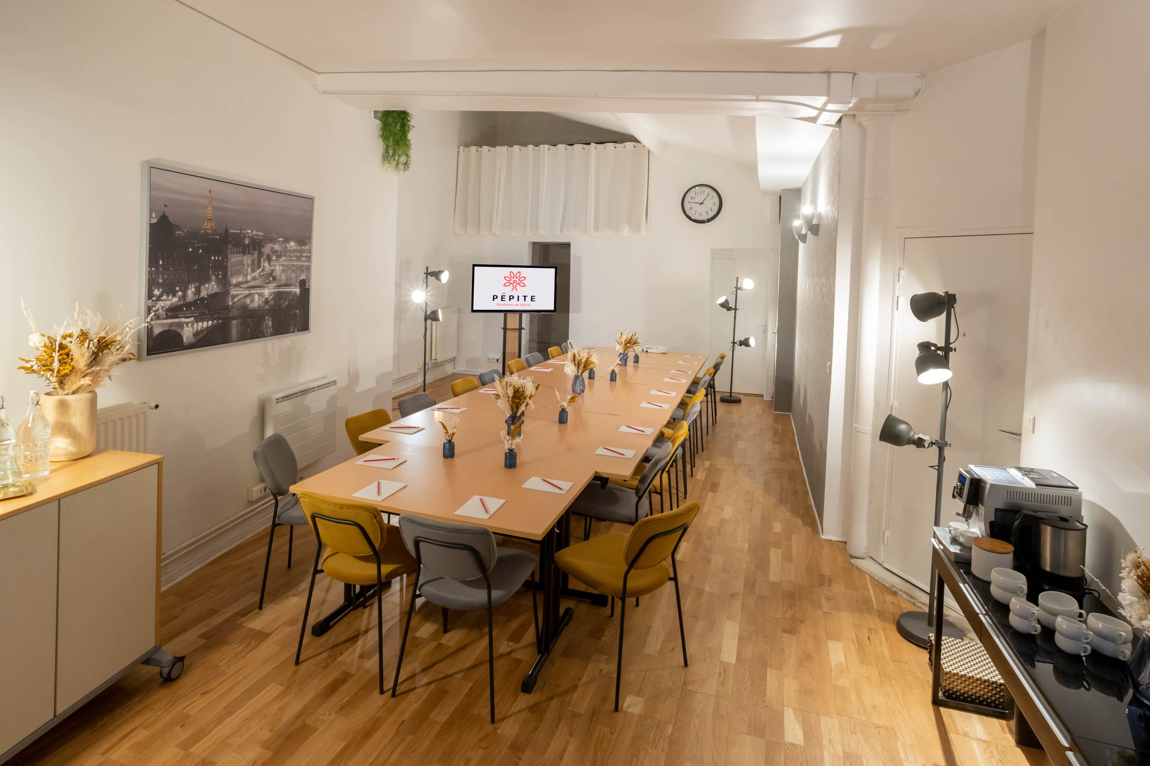 A long wooden table set for a meeting in a well-lit conference room with a wall clock, modern lamps, and decor.