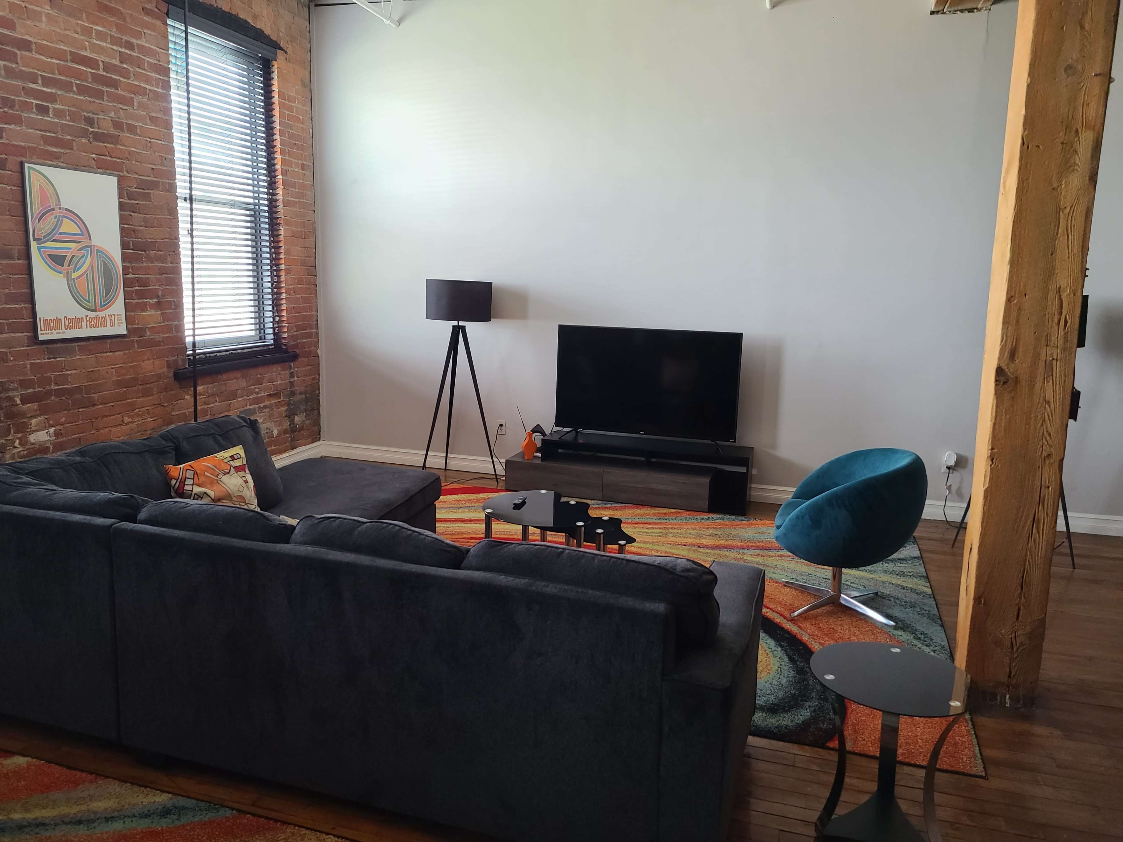 A modern living room featuring a dark sectional sofa, a large television on a stand, a blue accent chair, and a patterned area rug on wooden flooring.