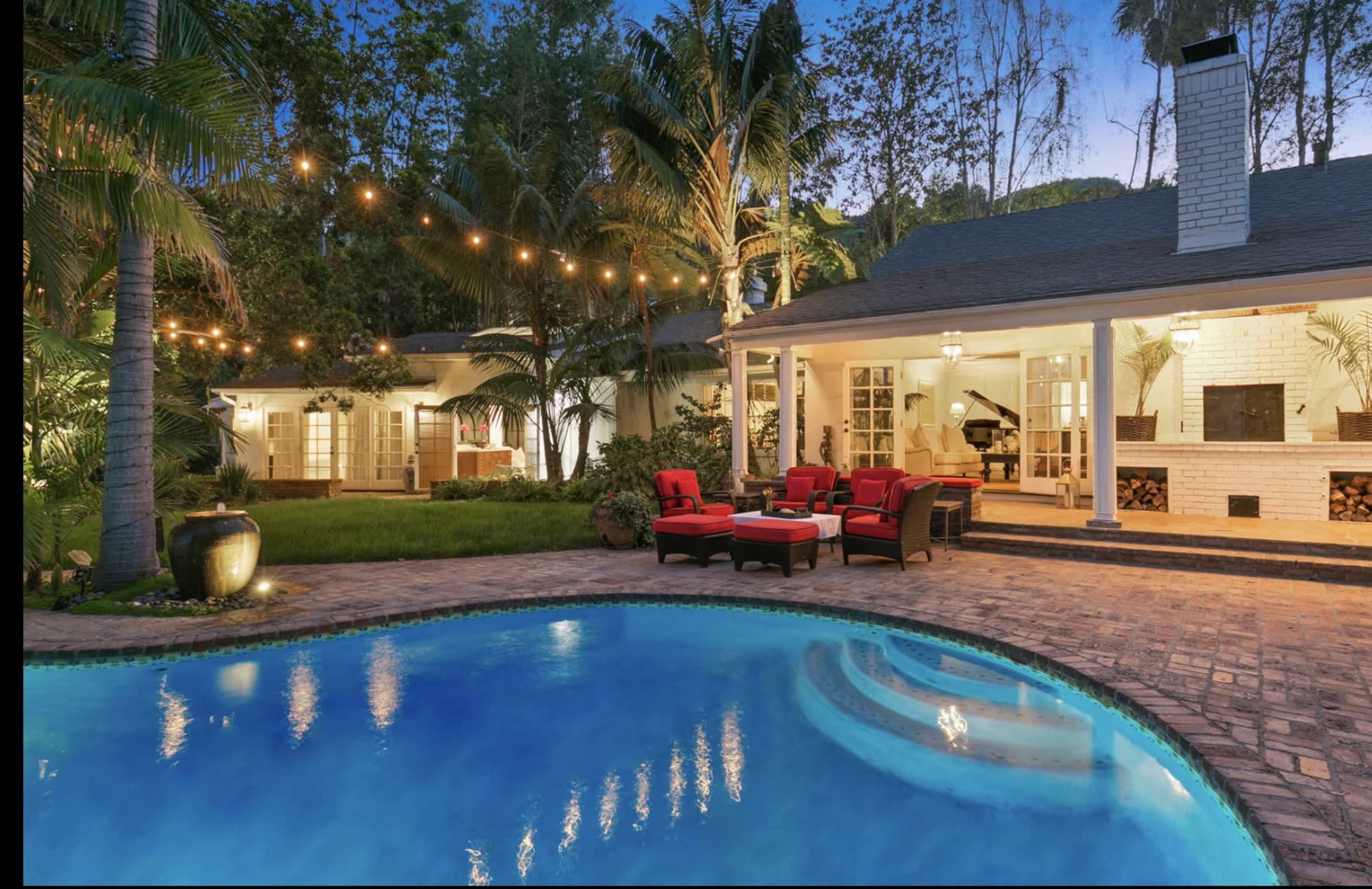 A well-lit backyard features a swimming pool surrounded by lounge chairs, with a house and outdoor seating area visible in the background.