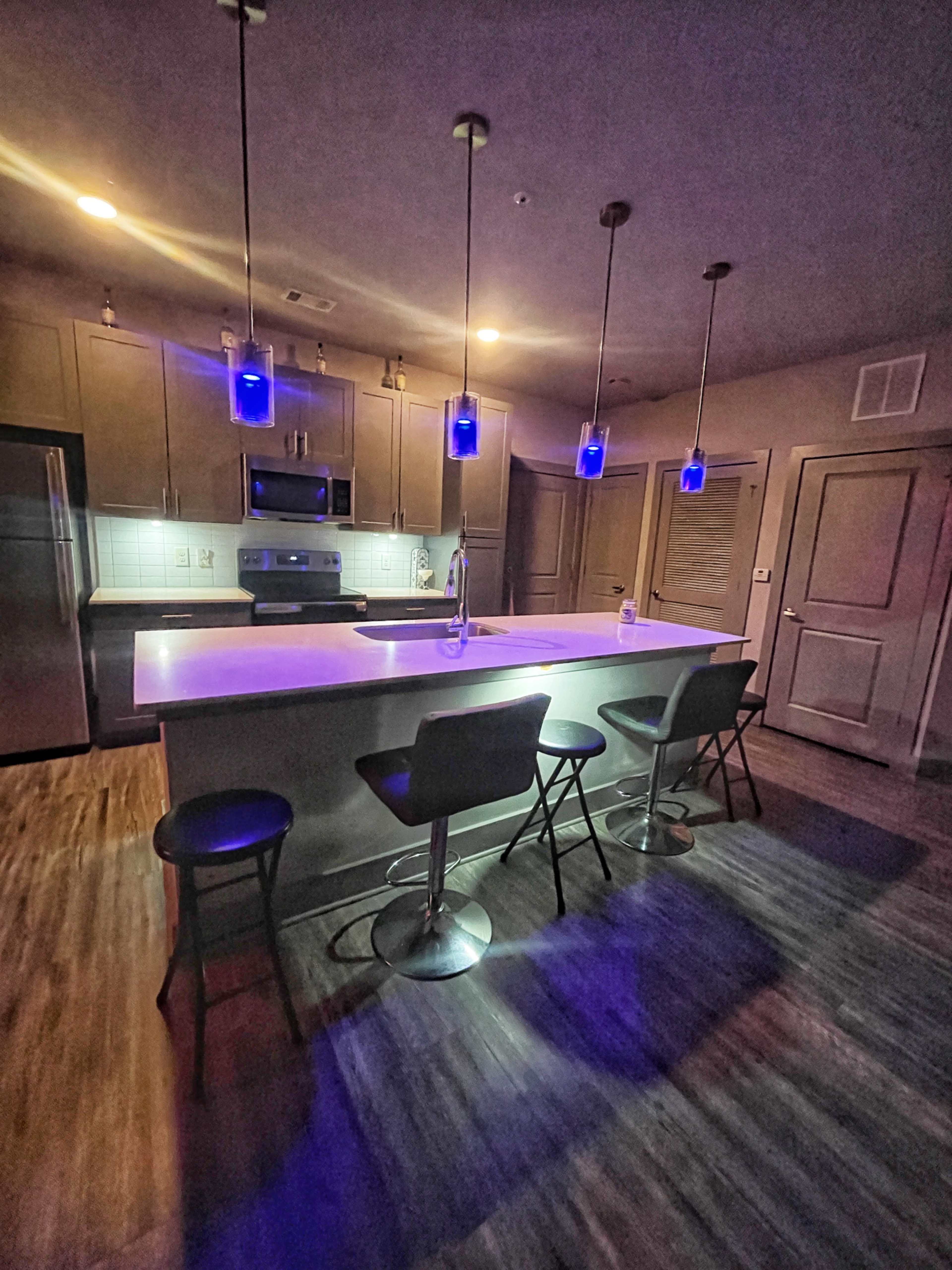 The kitchen features modern appliances, a central island with four high stools, and pendant lights illuminating the space with a blue glow.