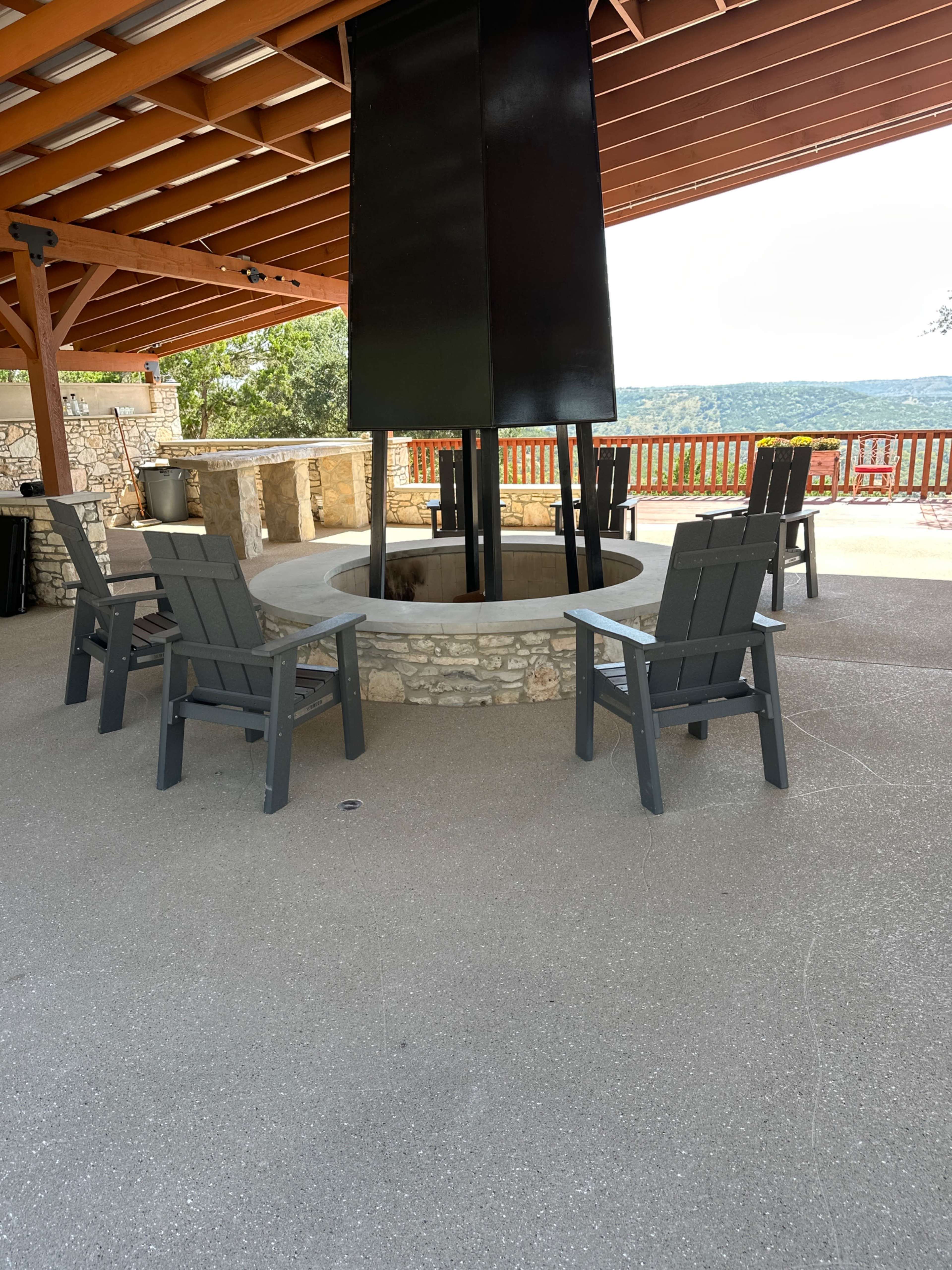 The image shows a circular stone fire pit surrounded by four adirondack chairs under a covered patio with a scenic view in the background.