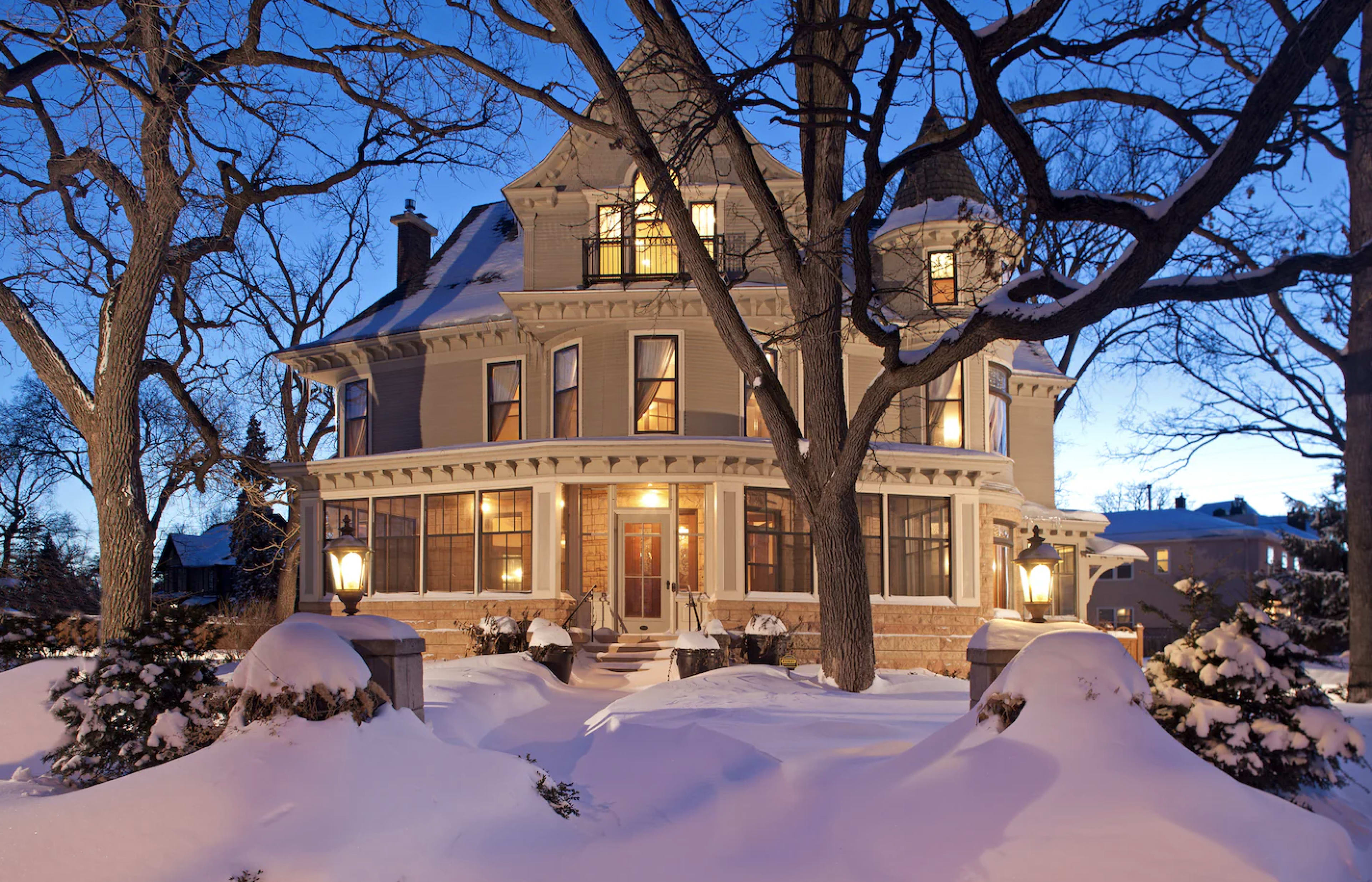 A large, historic house is surrounded by snow and illuminated by street lamps during twilight.