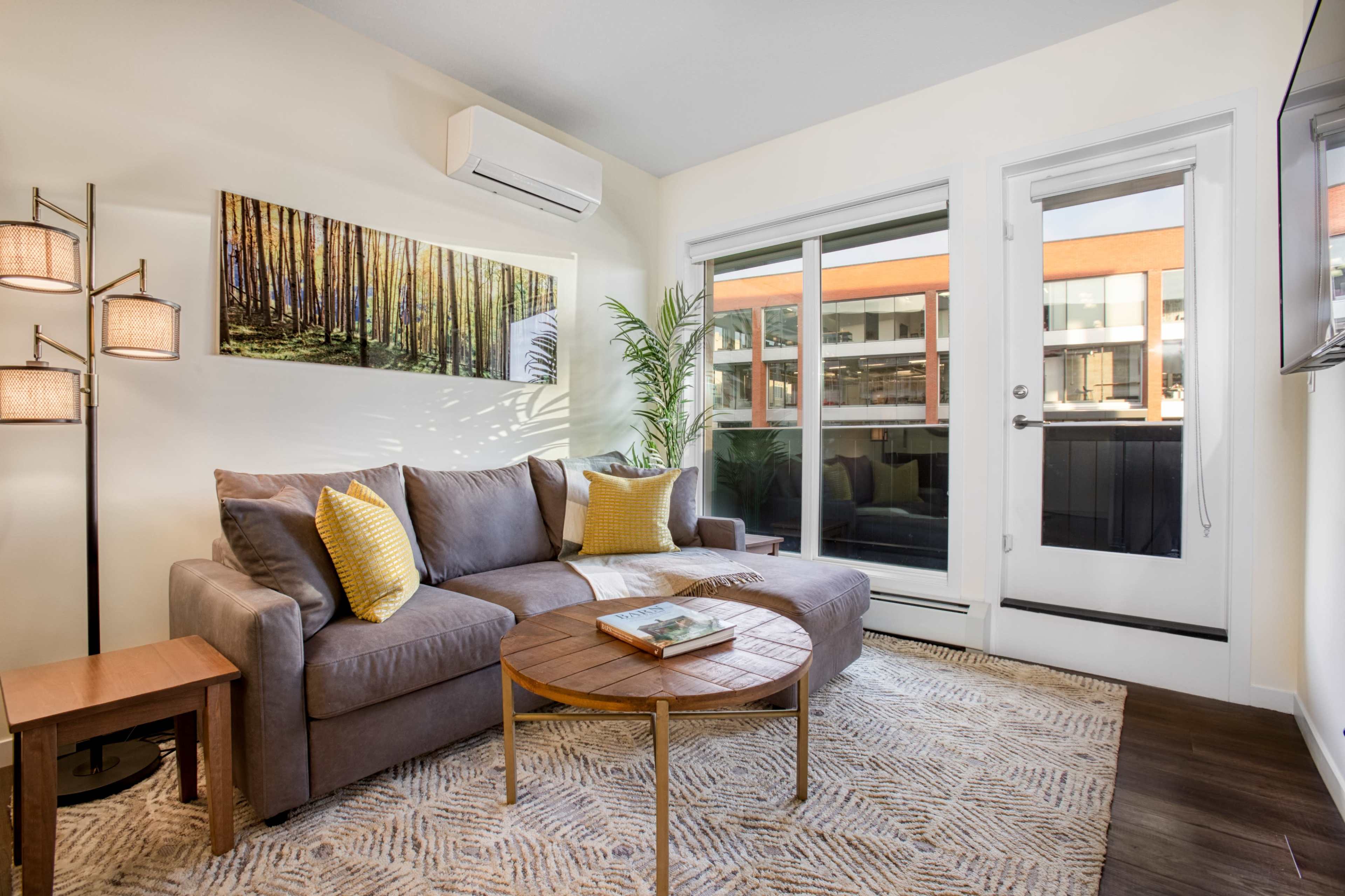 A cozy living room features a gray sectional sofa with yellow accent pillows, a round wooden coffee table, and large windows that overlook an outdoor patio.