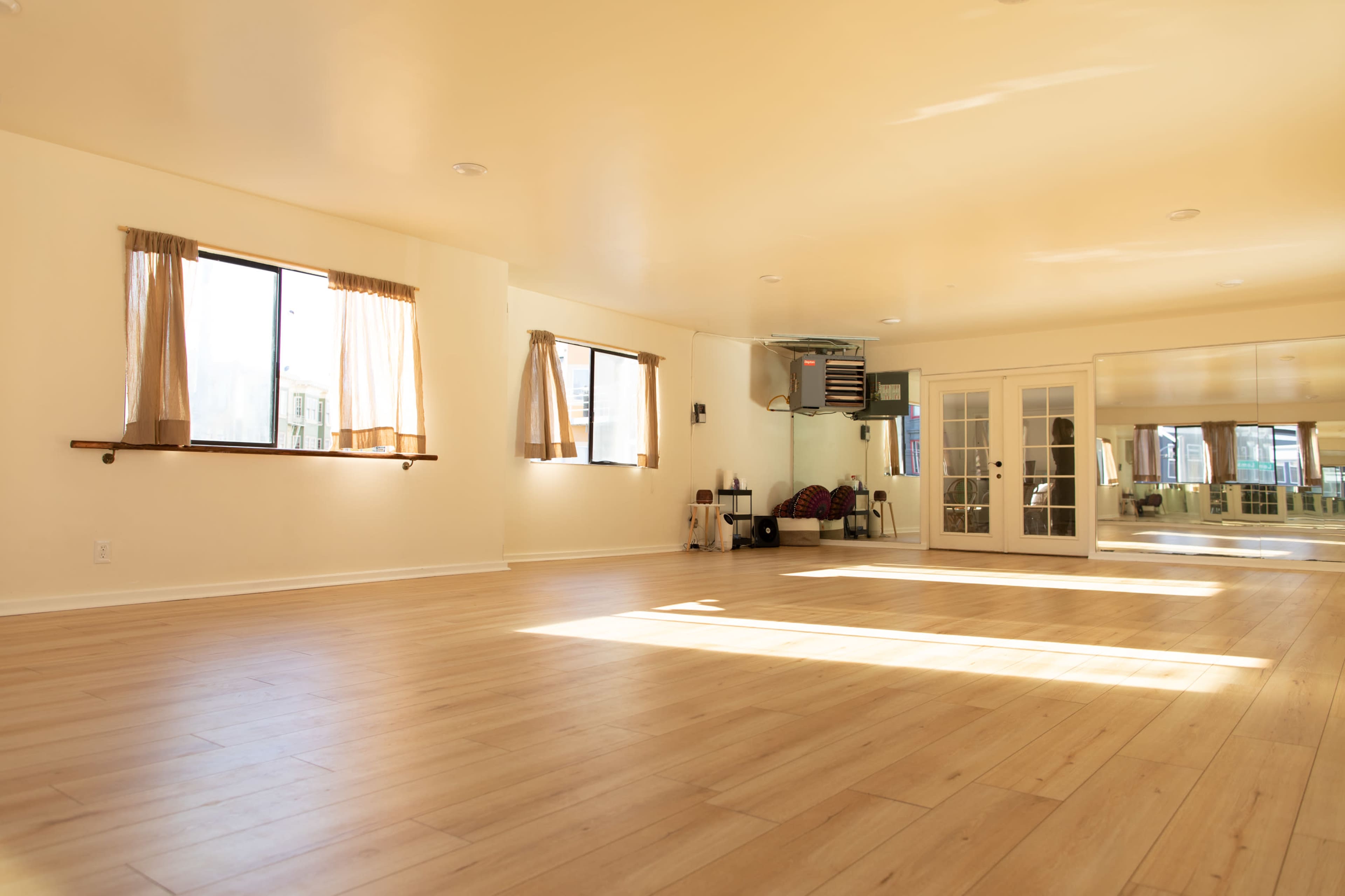 A spacious, well-lit room with wooden flooring, large windows, and mirrors on one wall.